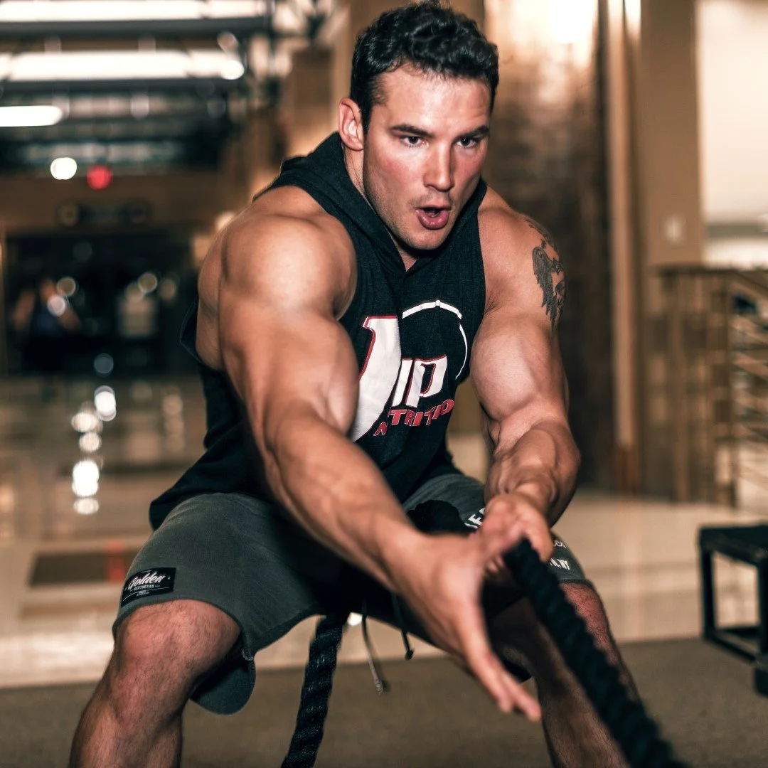A muscular man with short dark hair wearing a sleeveless black shirt and gray shorts is engaged in a battle rope workout inside a gym.