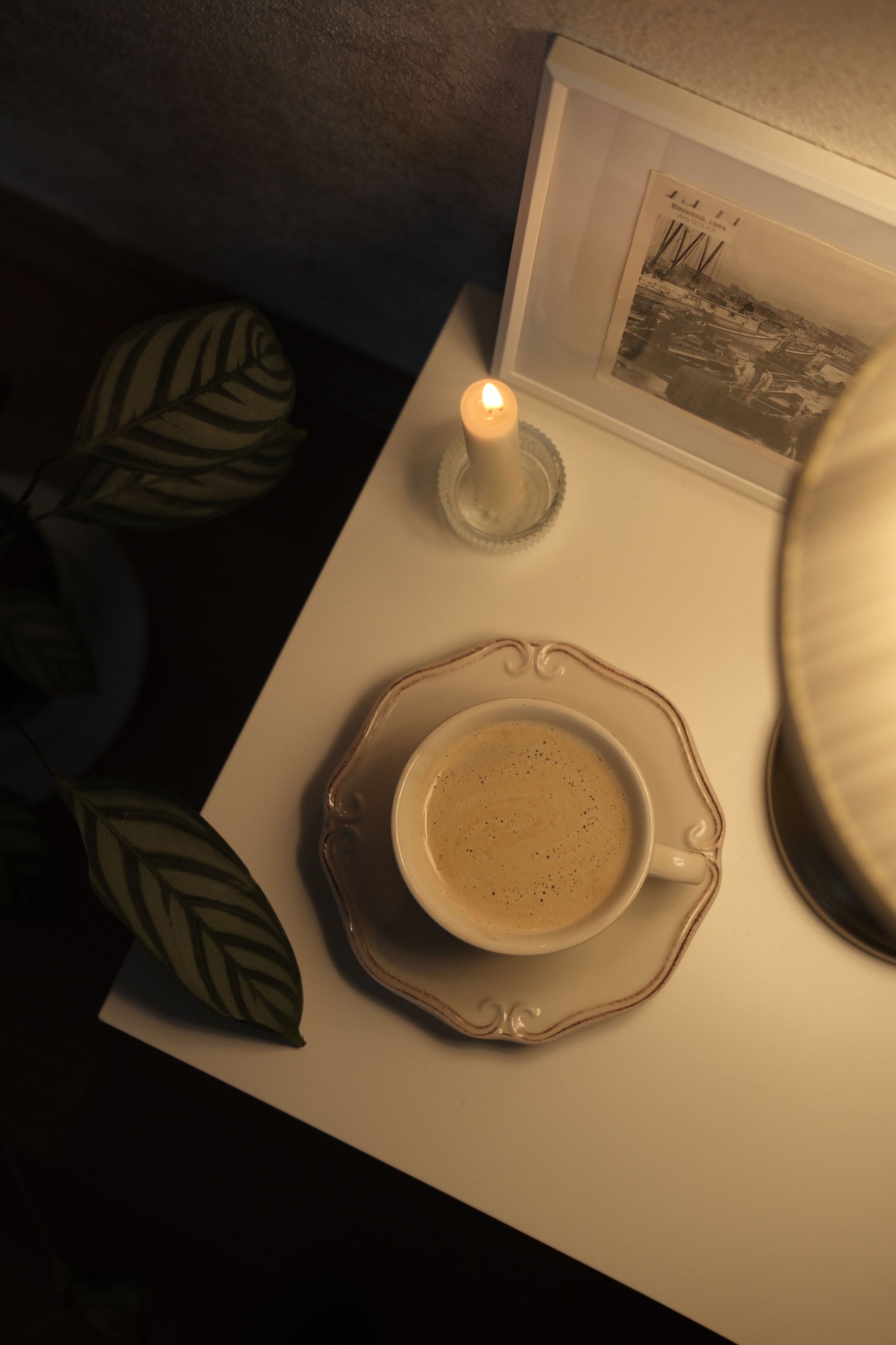 A lit candle in a glass holder, a framed black-and-white photograph of a harbor, a decorative teacup filled with coffee, and green leafy plants on a white surface.