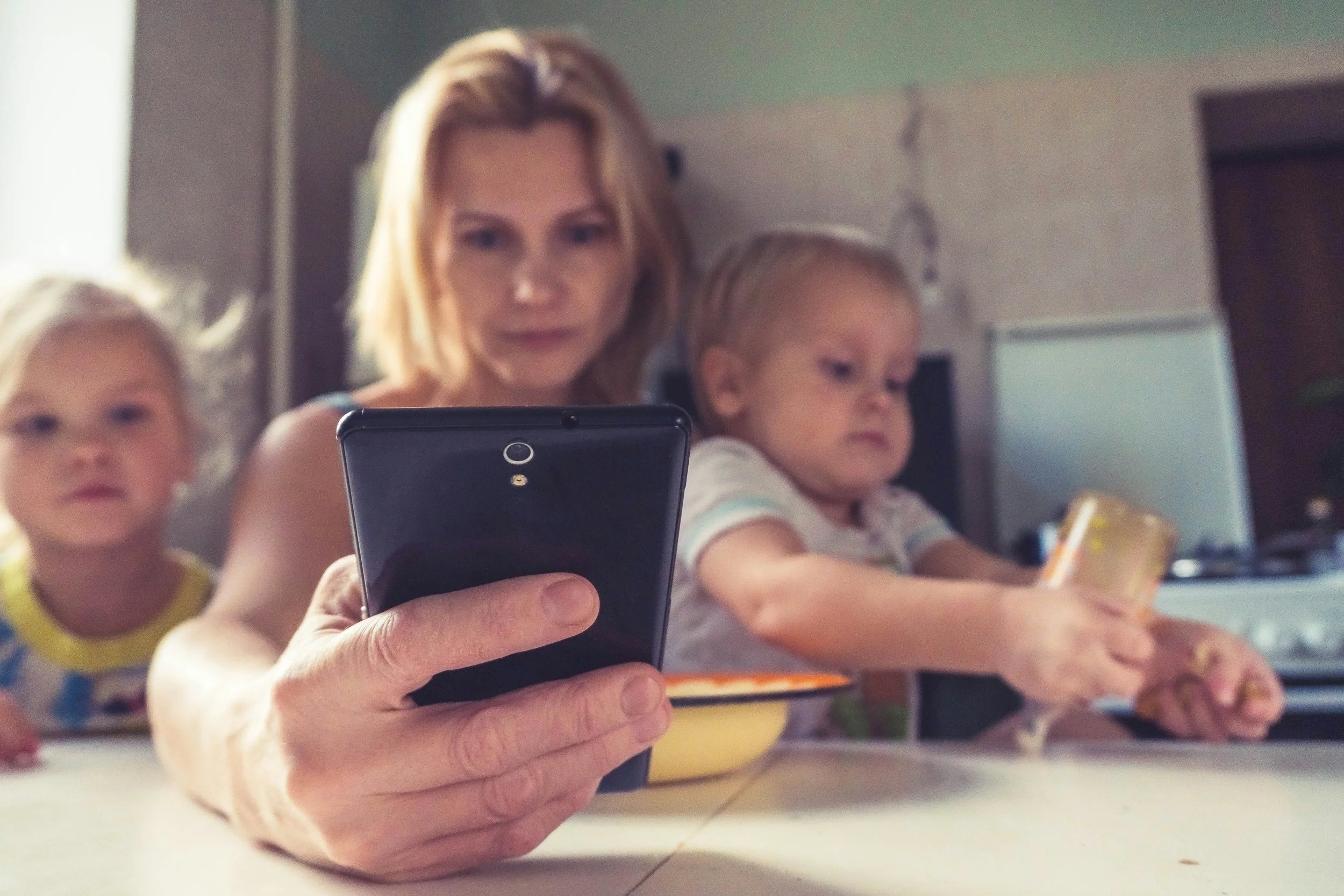 A woman and two children sitting at a kitchen table, with the woman holding a smartphone up to take a selfie, while the children are engaged with their food and a yellow cup.