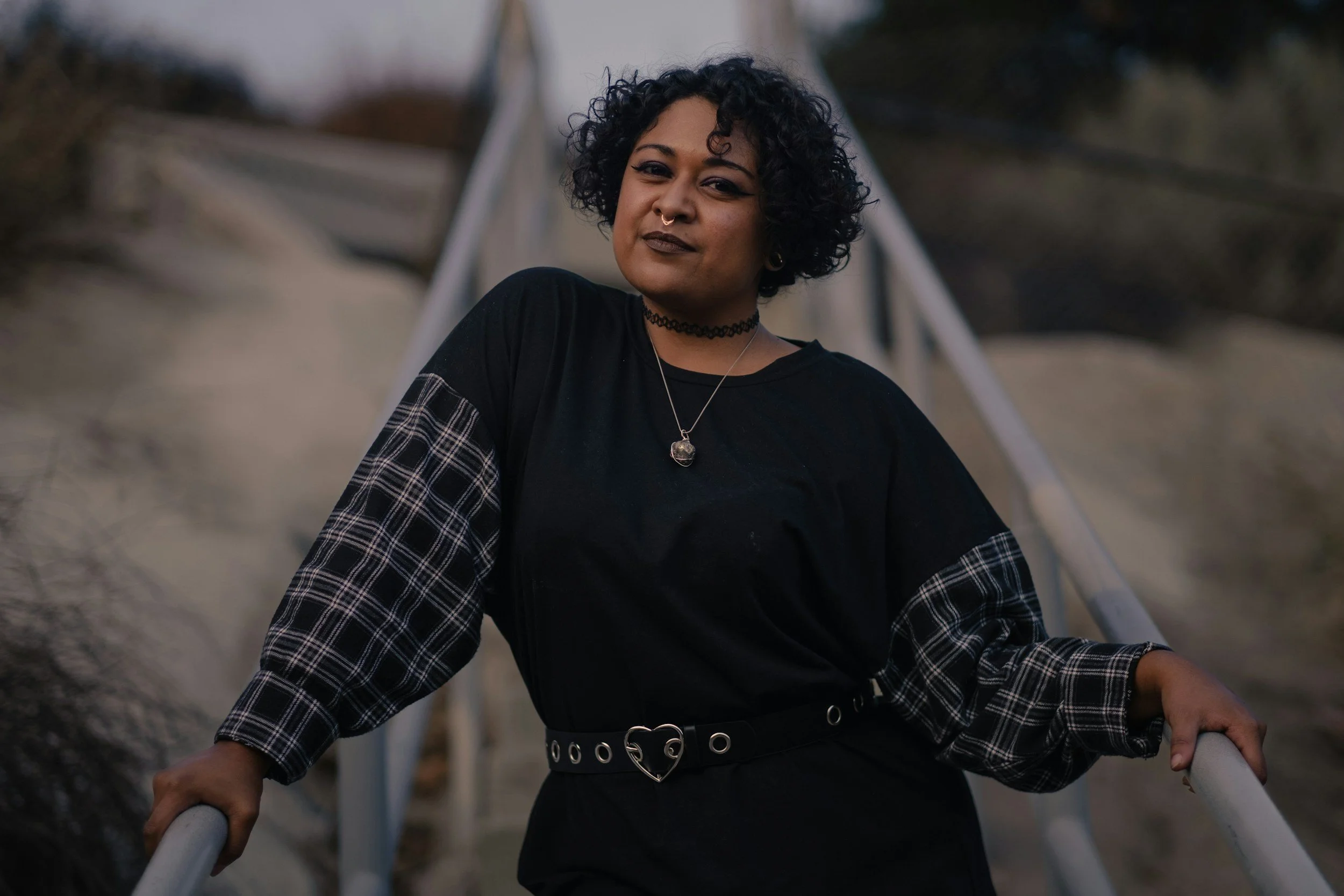 A woman with curly black hair wearing a black shirt with plaid sleeves, standing on a staircase outdoors, holding the railing with both hands, and looking at the camera.