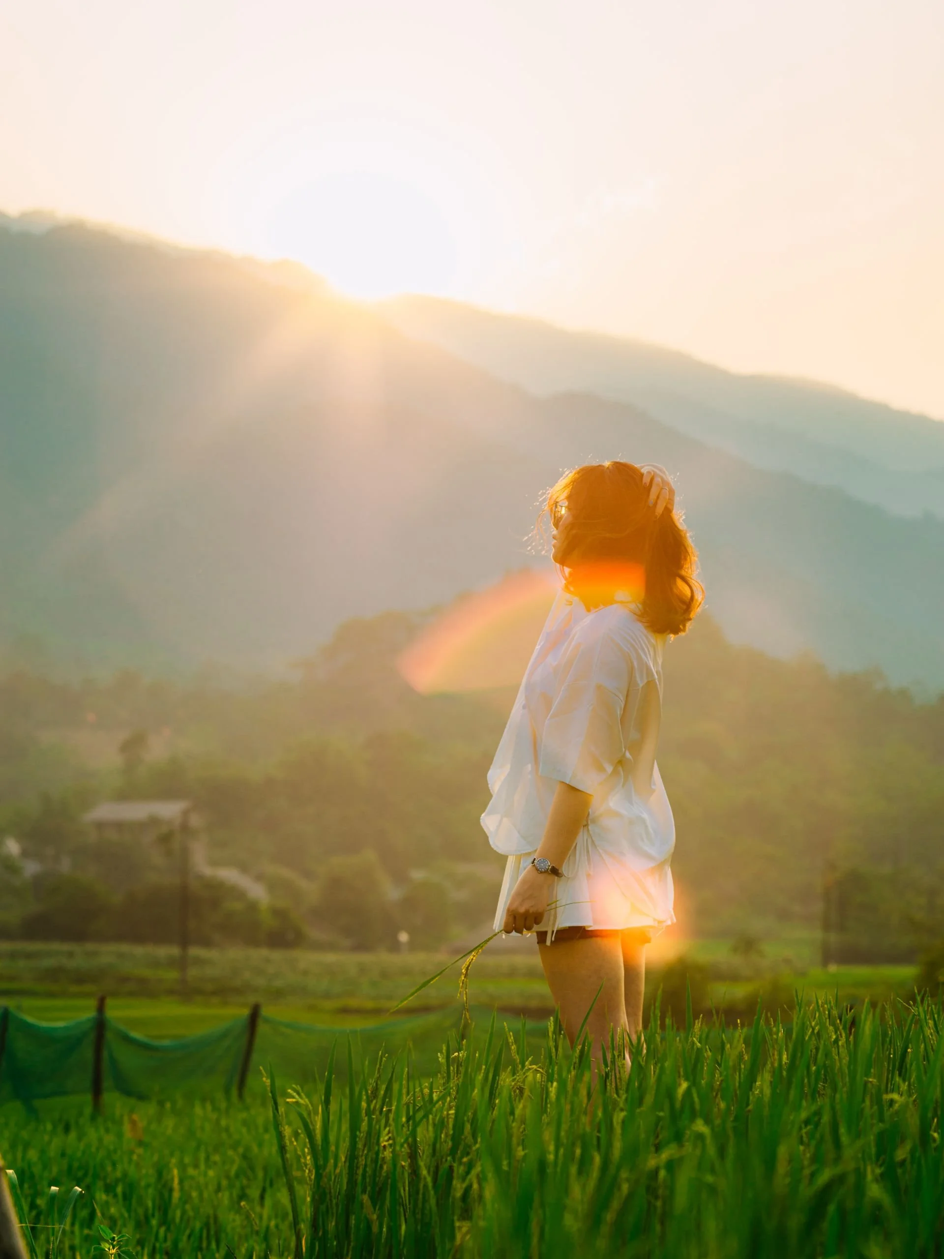 A woman standing in a lush green field during sunset, with mountains in the background and sunlight creating a warm glow around her.