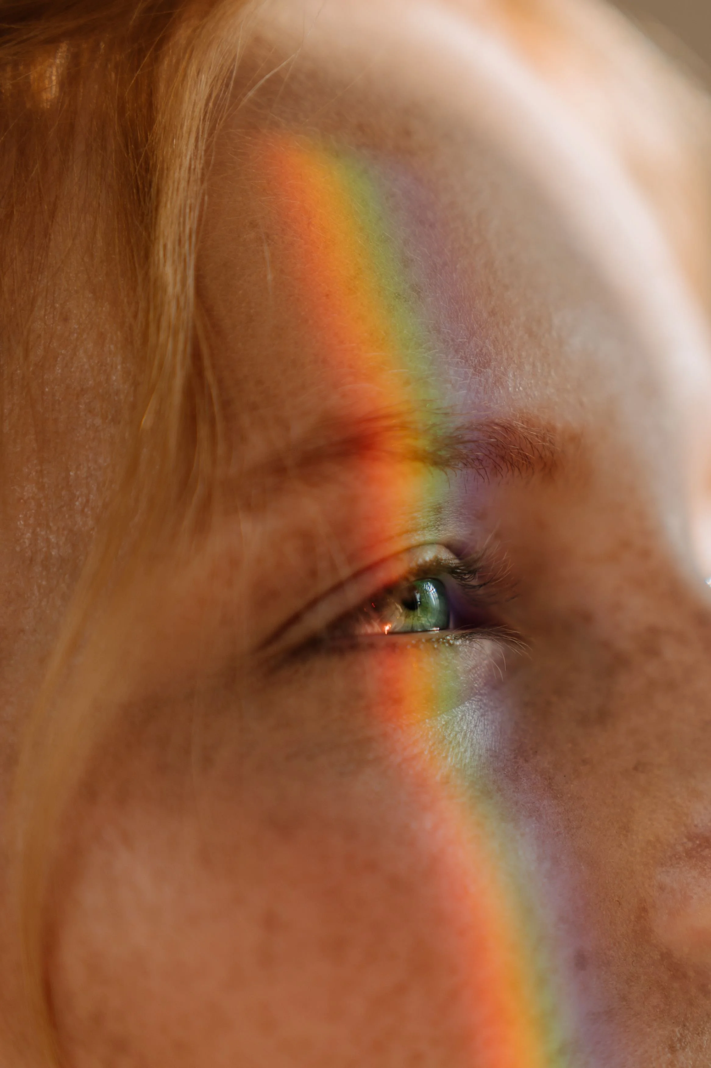 Close-up of a person's face with a rainbow reflection across the eye and cheek.