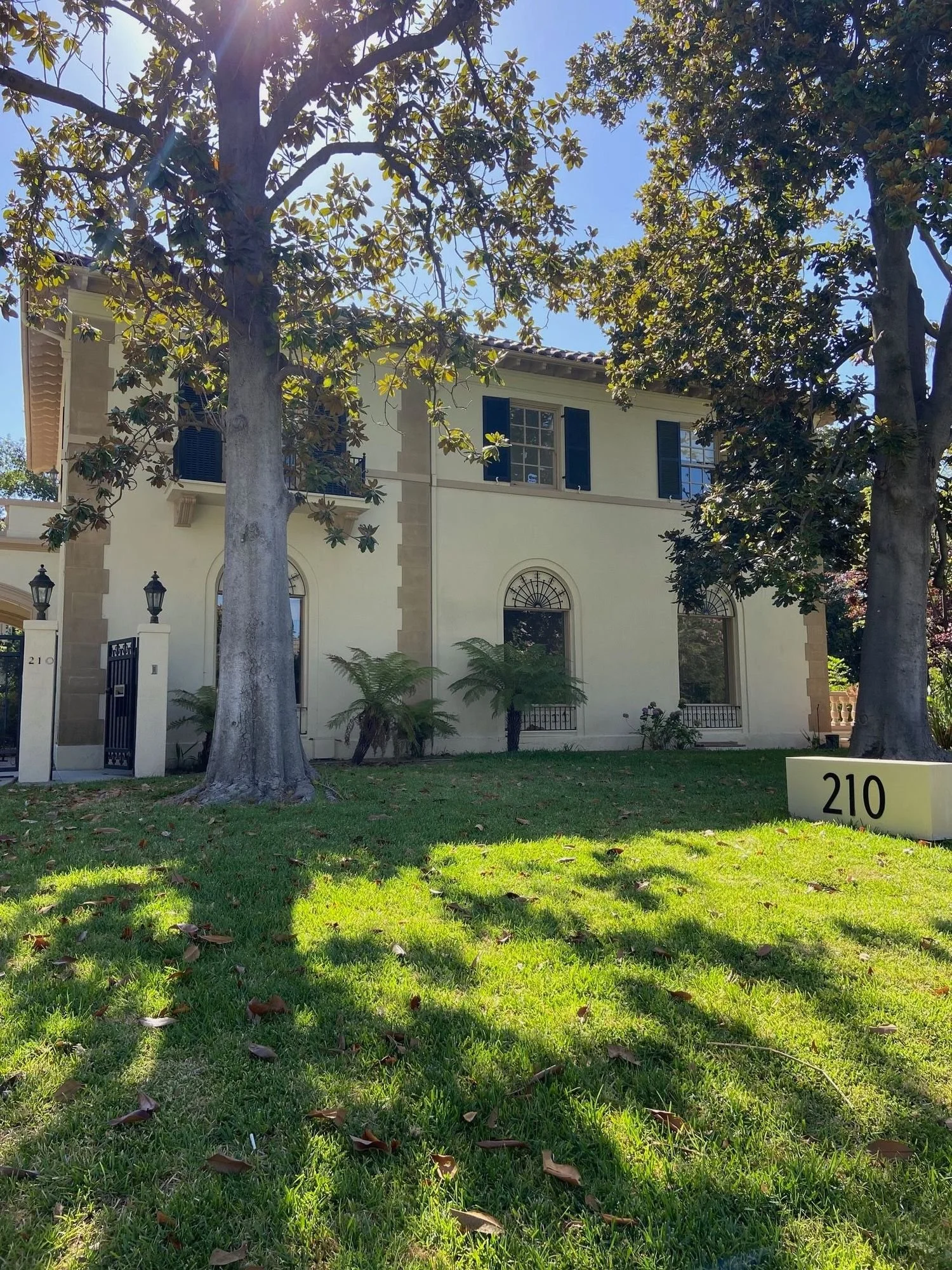 A two-story house with cream-colored walls and dark blue shutters, surrounded by large trees with green leaves. The house has arched windows and a wrought iron fence with gate near the front entrance. A white sign with black numbers '210' is on the lawn, which is green and shaded by trees. The sun is shining from the top left corner, creating bright light and shadows.
