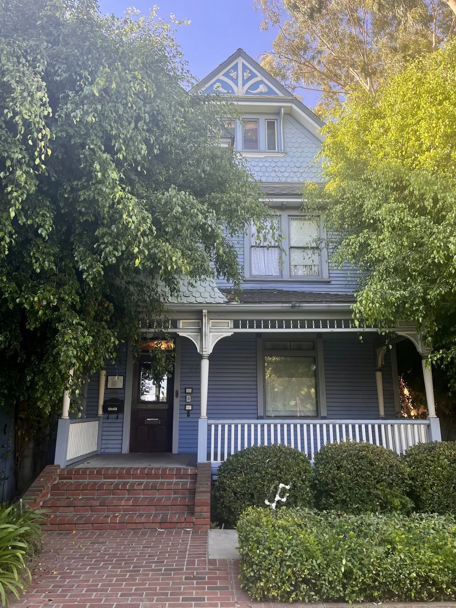 A blue Victorian house with white trim, surrounded by green trees and bushes, with a brick pathway and steps leading to the front door.