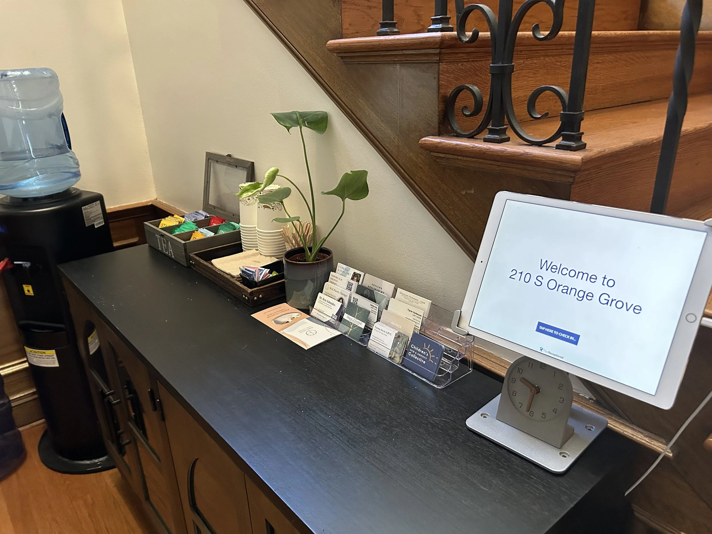 A reception area with a black countertop, a water cooler, a tray of tea packets, a potted plant, and a digital display screen showing the address 210 S Orange Grove.