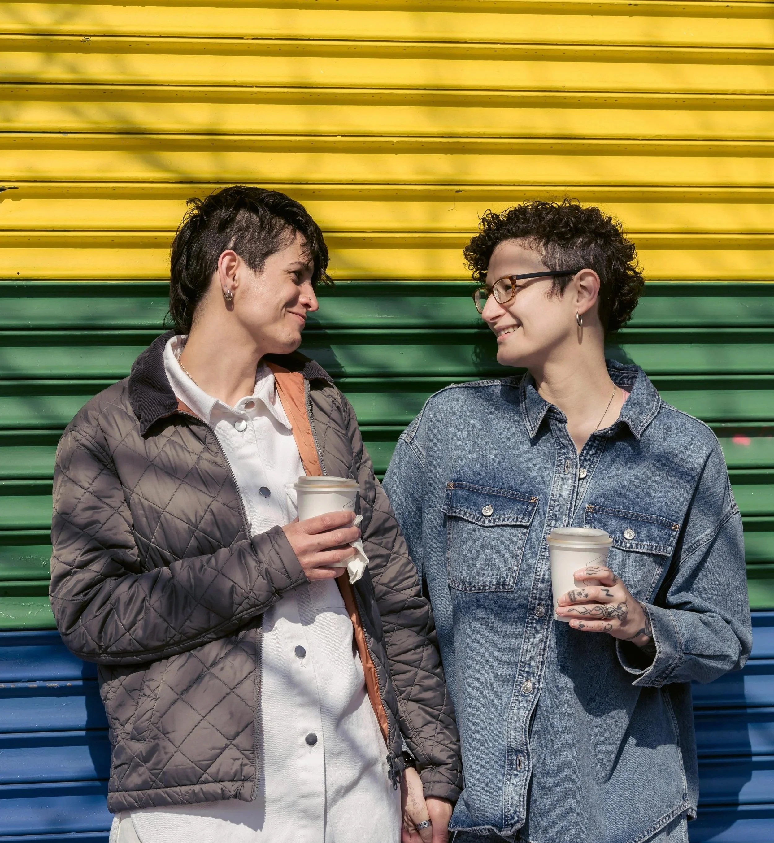 Two women with short, curly and mohawk hairstyles, smiling at each other, holding coffee cups, standing in front of a colorful, striped metal wall.