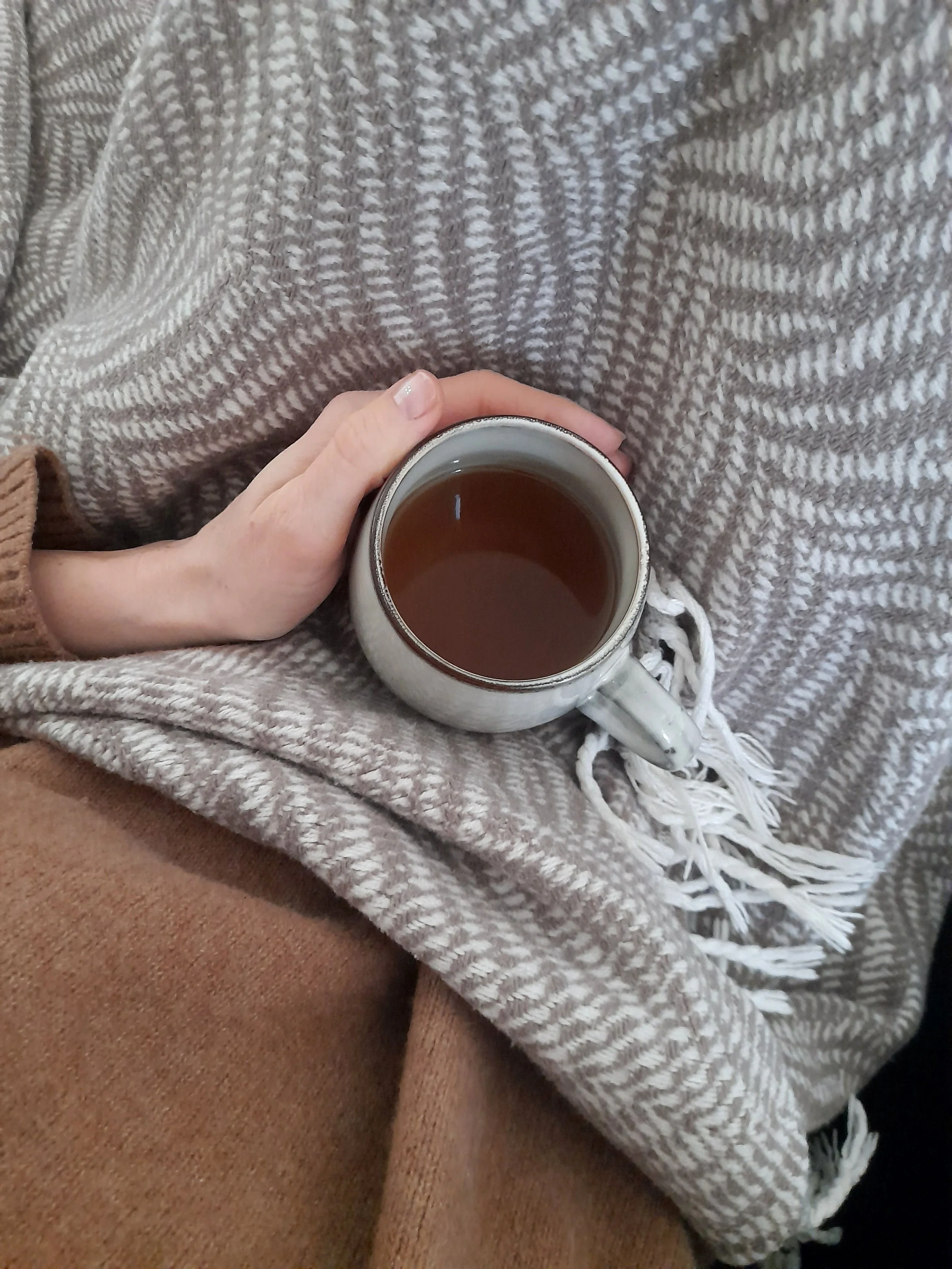 A person holding a coffee mug with a dark beverage, sitting on a brown and beige textured blanket. The person's hand is visible with neatly trimmed nails and a light-colored nail polish. The blanket has a checkered or woven pattern.