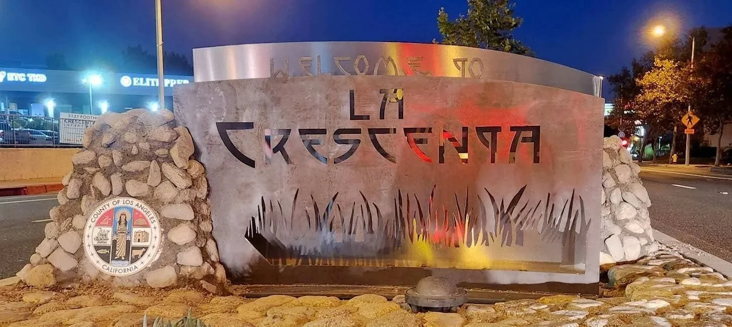 Street view of the entrance sign to La Crescenta, California, with stone and metal design, illuminated at night, with trees and streetlights in the background.