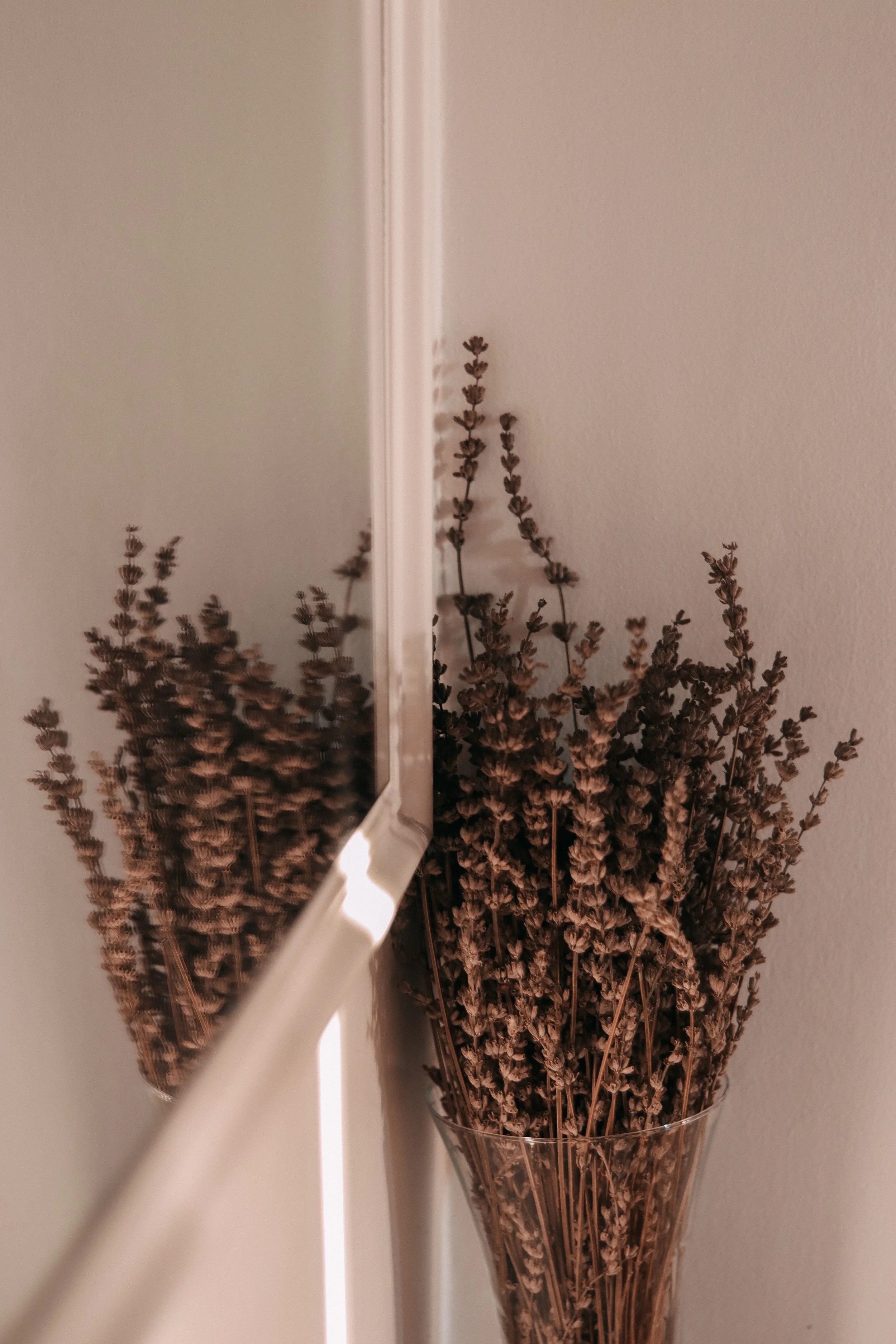 A close-up of a bouquet of dried lavender flowers in a clear glass vase, with a white wall in the background, partially covered by a white lighting strip.