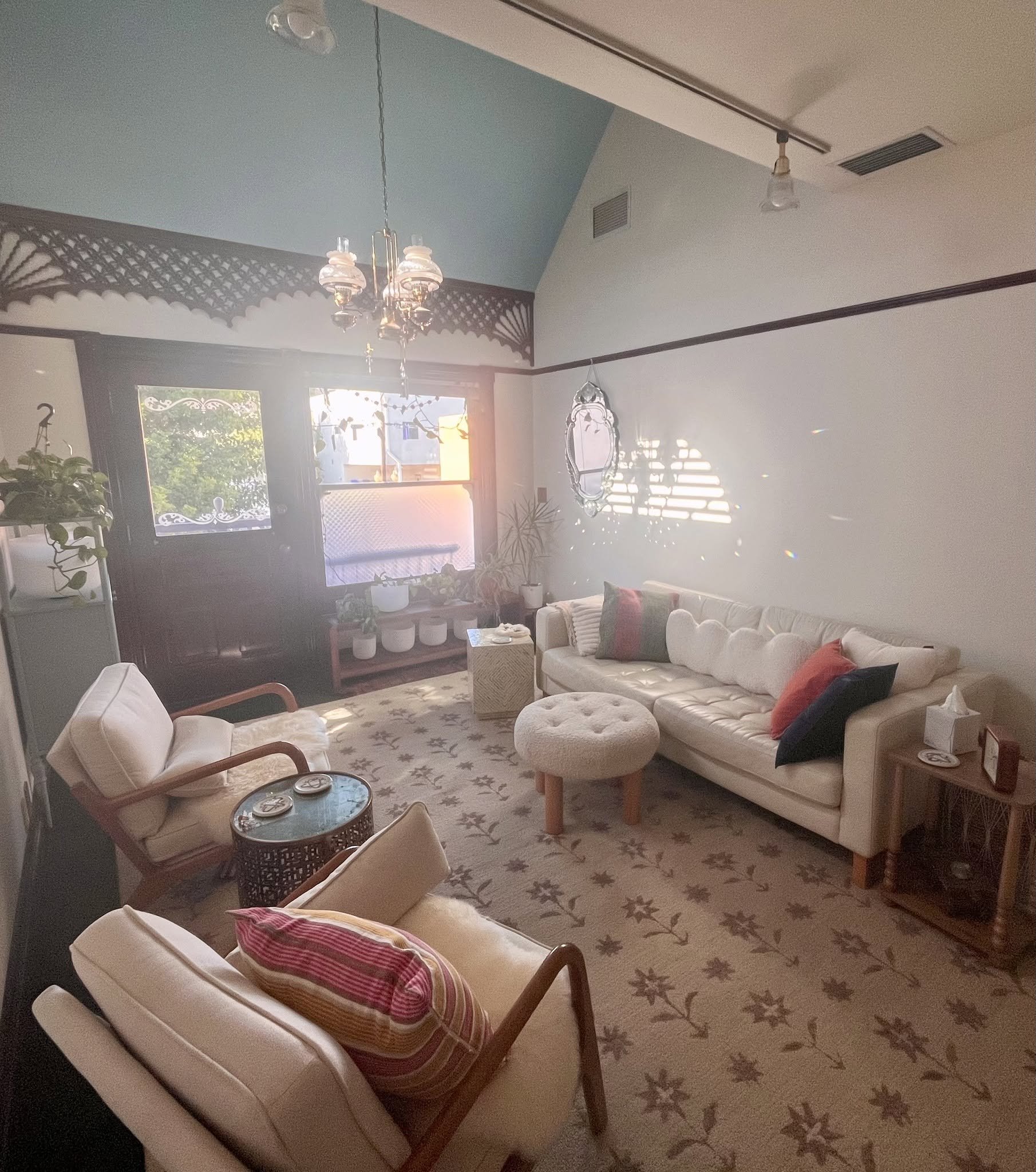 Living room with beige couch, armchair, and a patterned rug. A window with sunlight, plants, and decorative mirror on the wall. Ceiling lights and air vents visible.