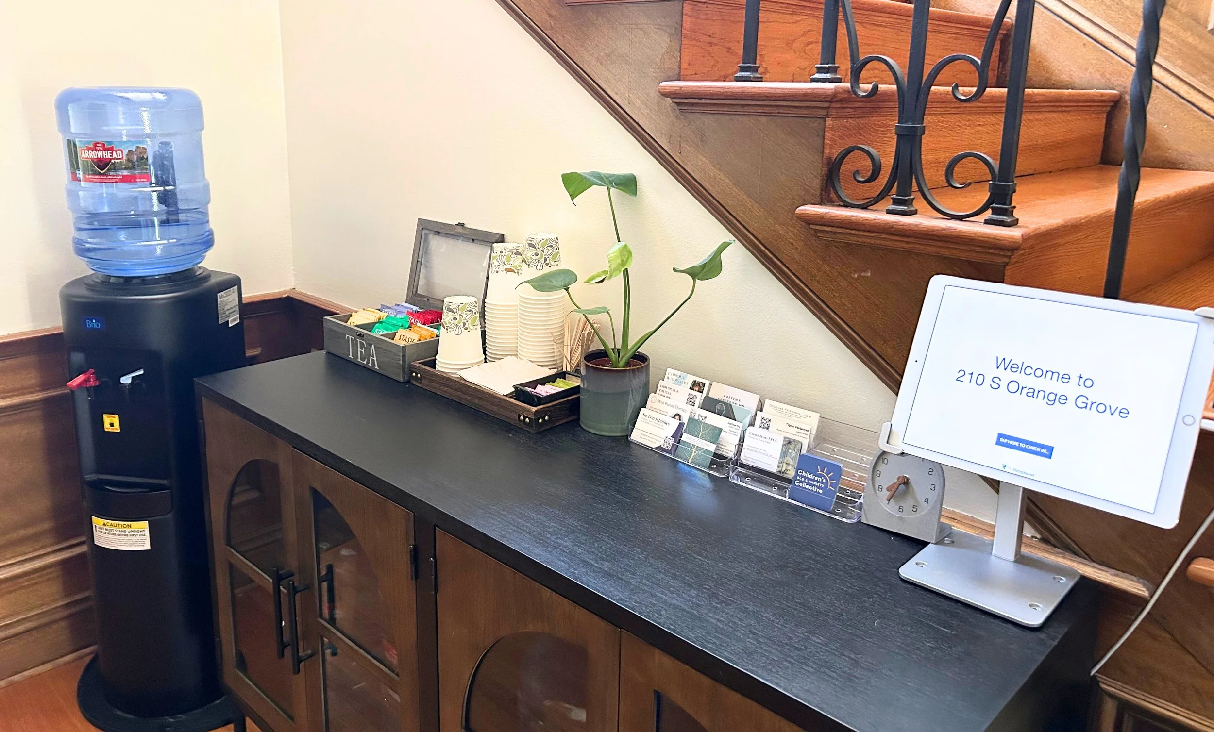 Coffee station with water dispenser, cups, tea boxes, and a welcome sign for 210 S Orange Grove.