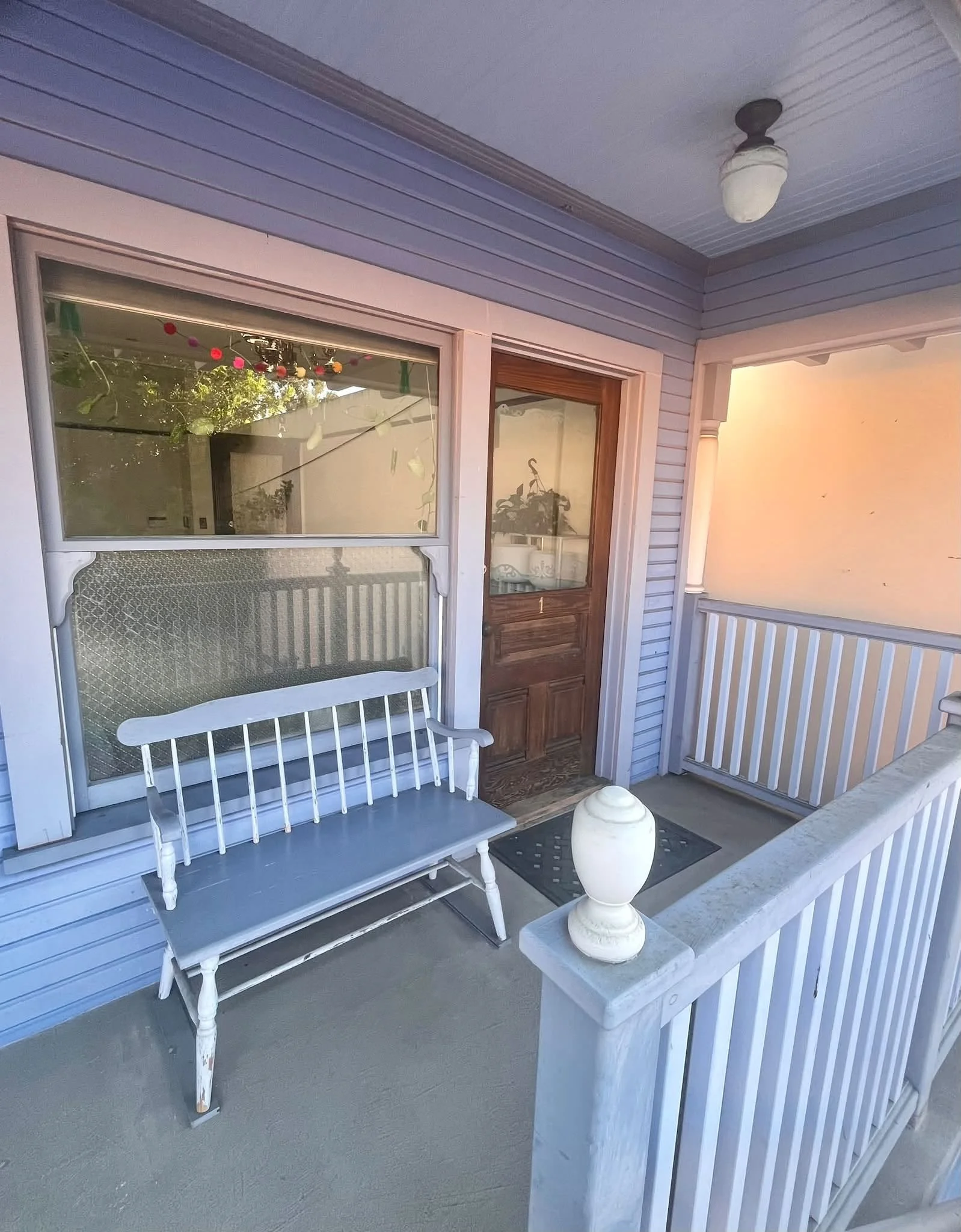 A balcony porch with a white wooden bench, a decorative vase on a small table, a window with a reflection, and a door, with a railing in front and a ceiling light fixture, during sunset.