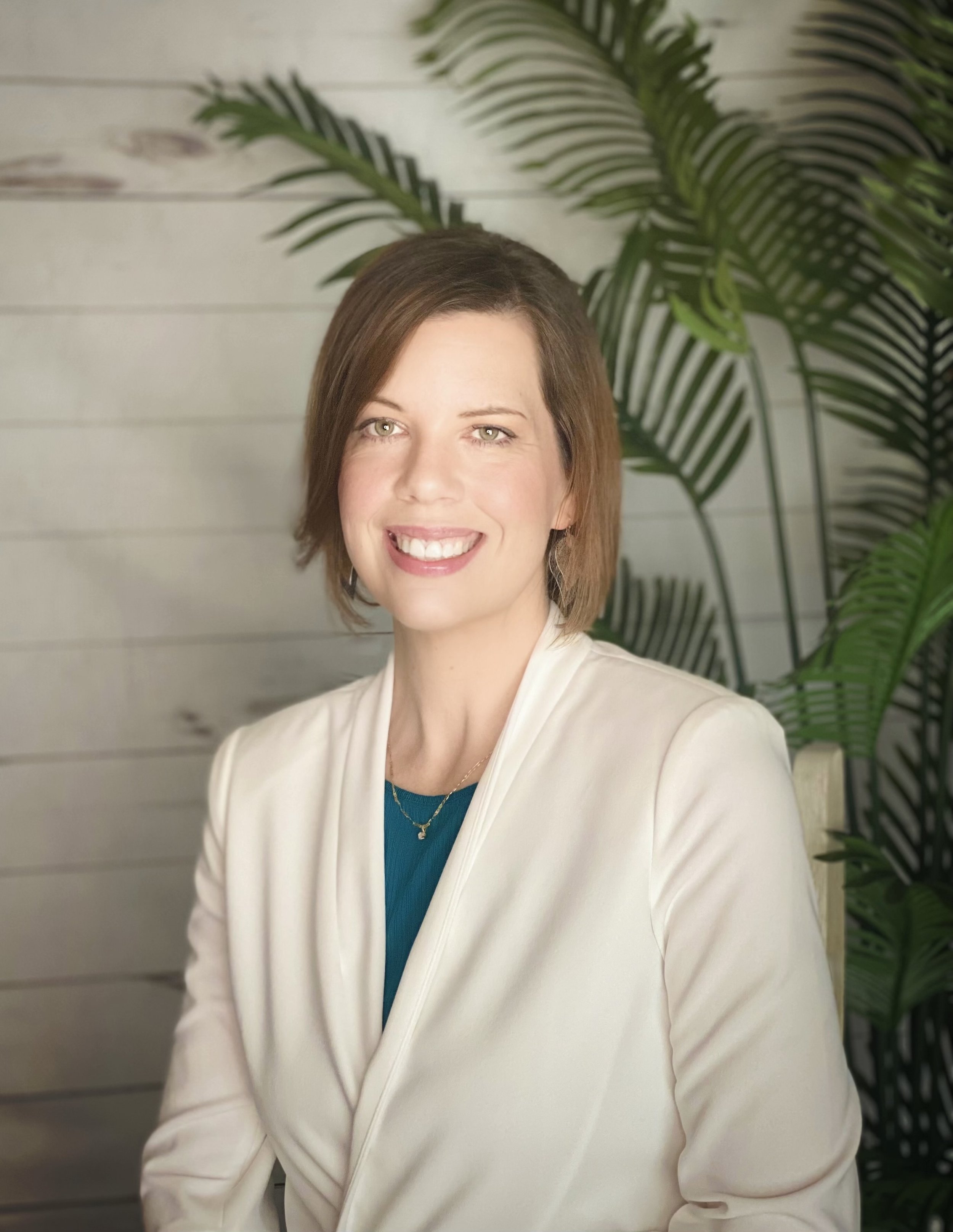 A woman with shoulder-length brown hair smiling, wearing a white blazer over a blue top, standing in front of green plant leaves and a light-colored wooden wall.