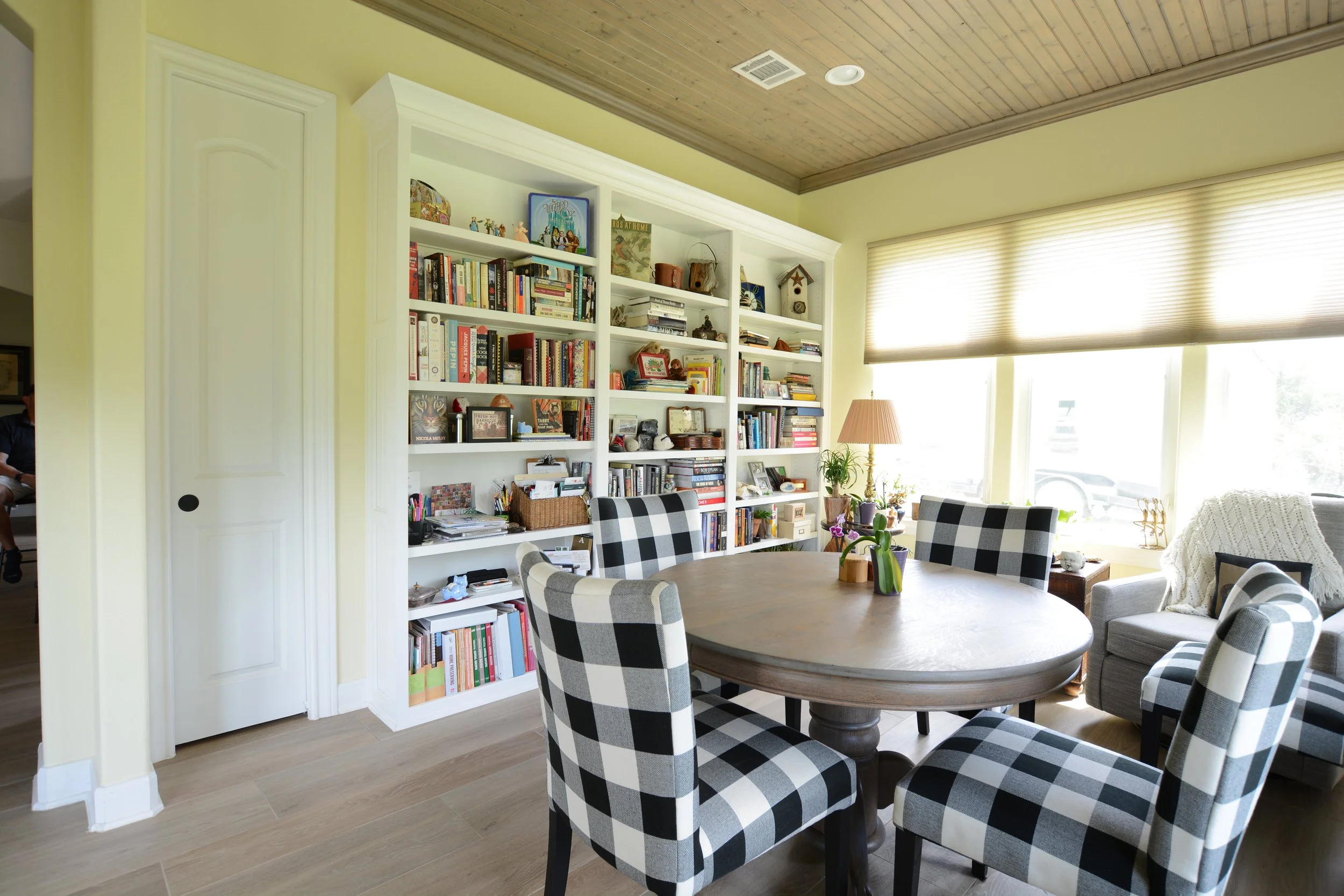 A cozy dining area with a wooden table surrounded by black and white plaid chairs, a large bookshelf filled with books and decorative items, a window with blinds letting in natural light, a potted plant, and an armchair with a white knitted throw.