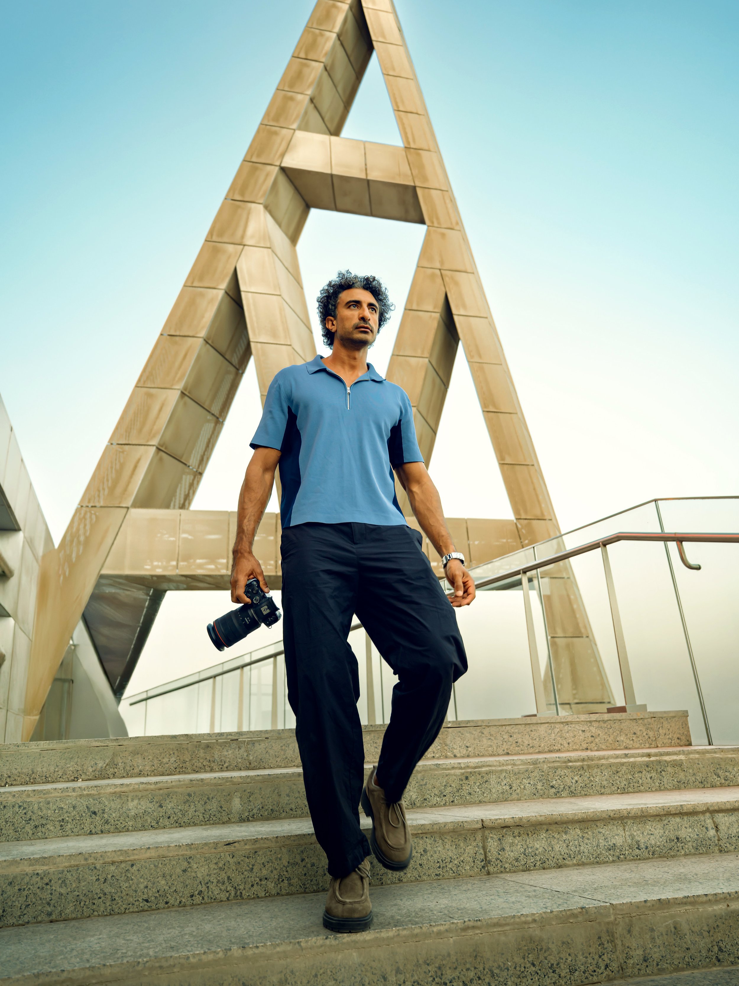 A man with curly hair dressed in a blue polo shirt and black pants walking down a staircase outdoors, holding a camera in his right hand. Behind him is a modern architectural structure with wooden panels and geometric design.