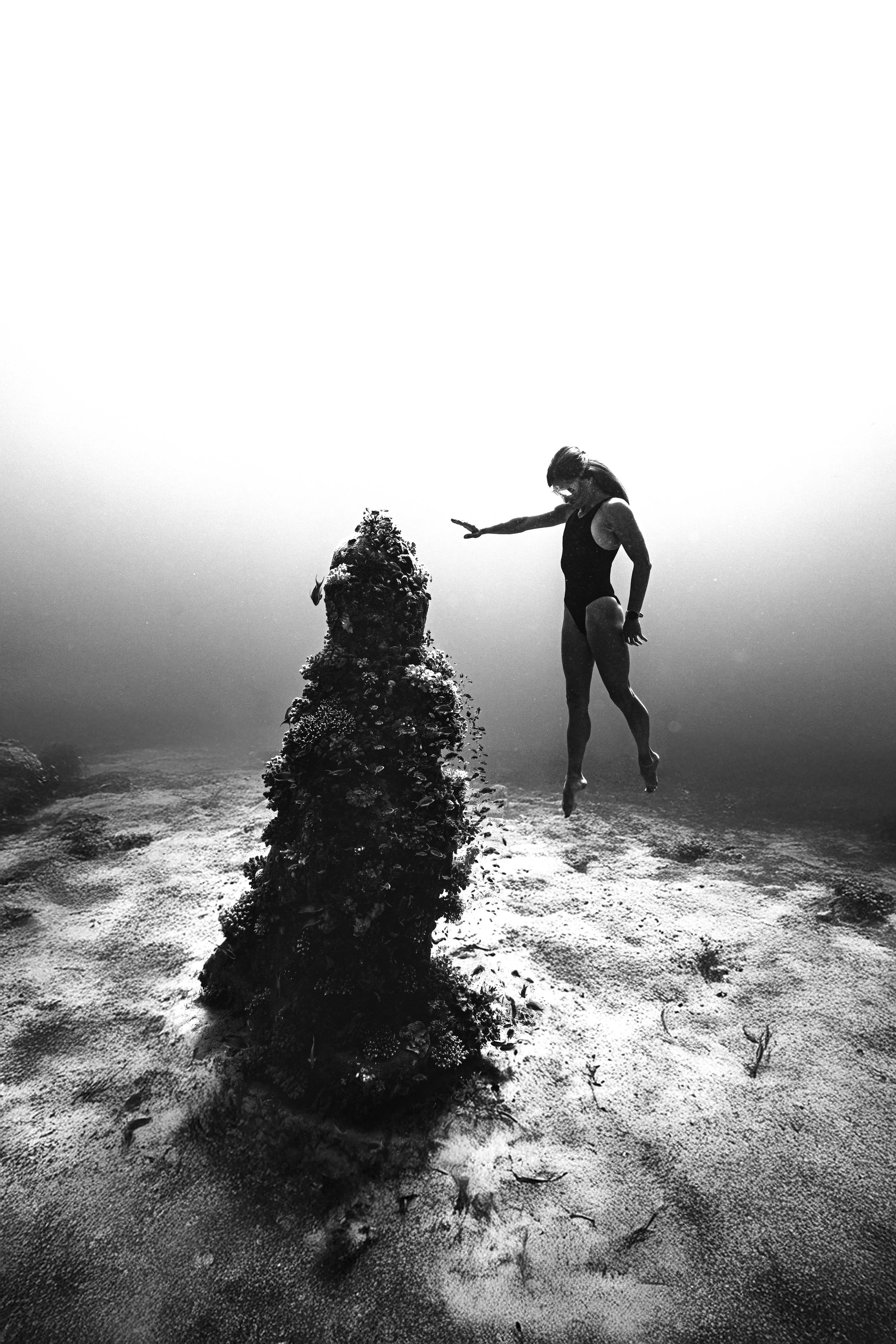 A woman in a swimsuit is jumping over a large rock covered with barnacles and marine growth in shallow water, with a bright sky in the background.
