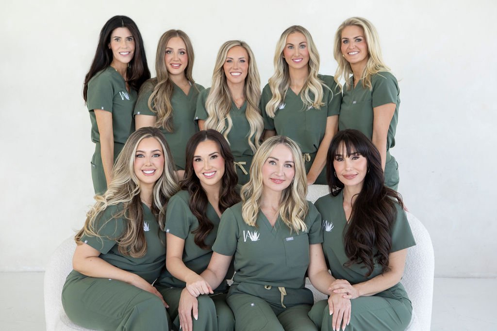 Group of nine women dressed in medical scrubs, smiling, posing together in a studio setting.