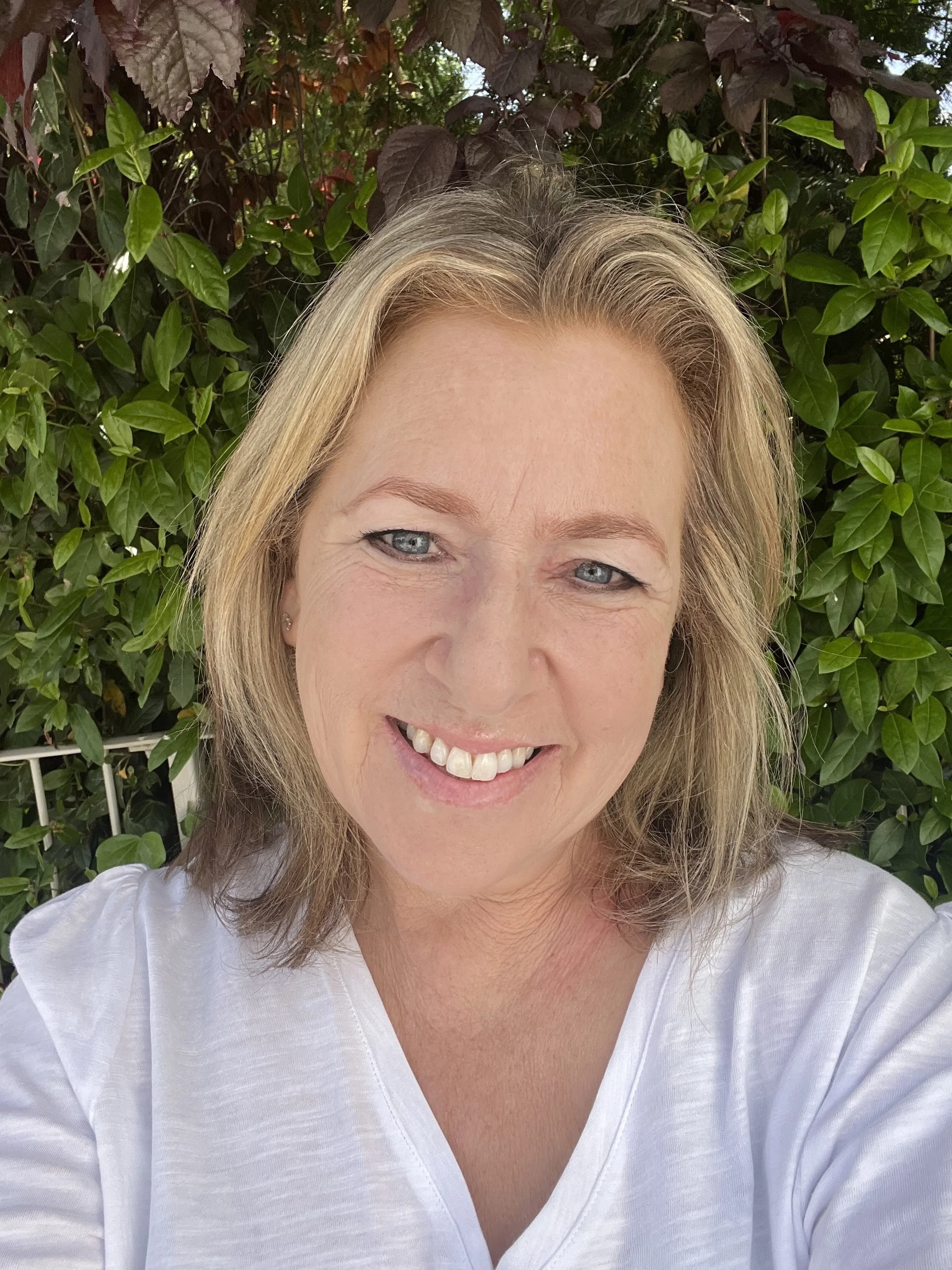Andee is a white woman with blond shoulder length hair. The photo is taken outside with greenery in the background and she is smiling at the camera.