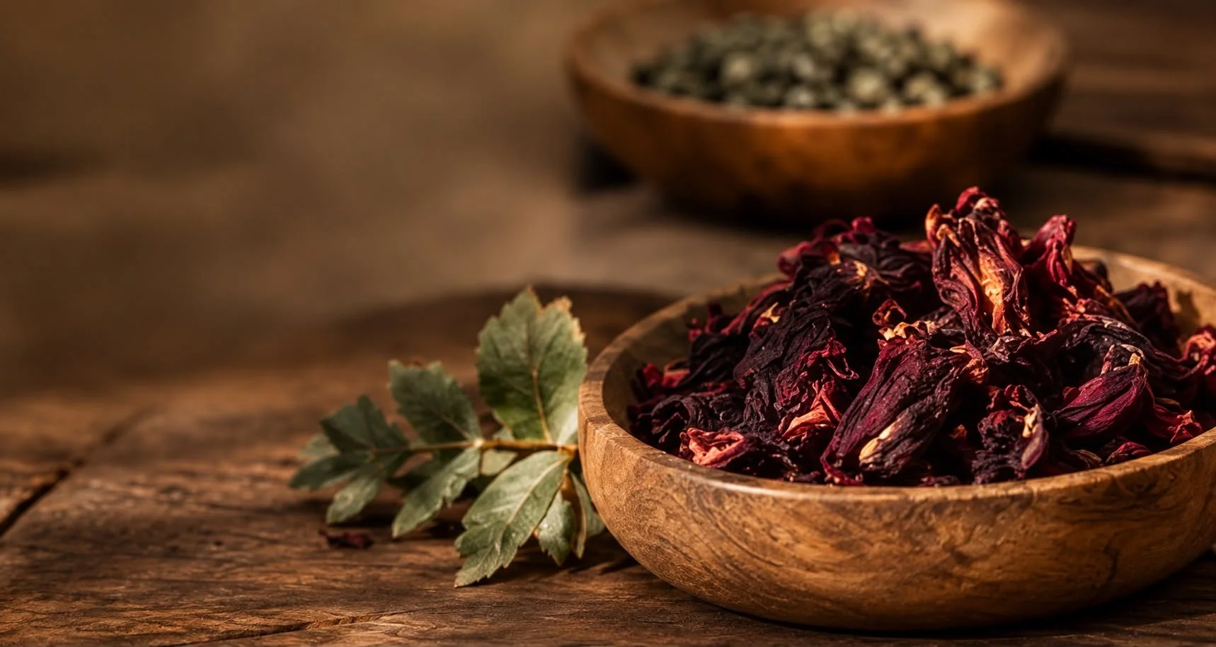 Tasses de hibiscus séché dans des bols en bois, avec des feuilles de géranium posées à côté, sur une surface en bois.