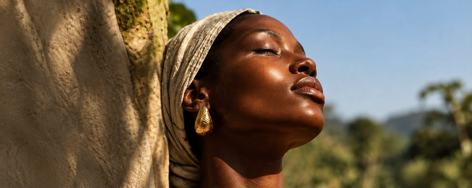 Femme noire portant un foulard clair et une boucles d'oreilles dorée, regardant vers le ciel avec une expression de sérénité, en plein air