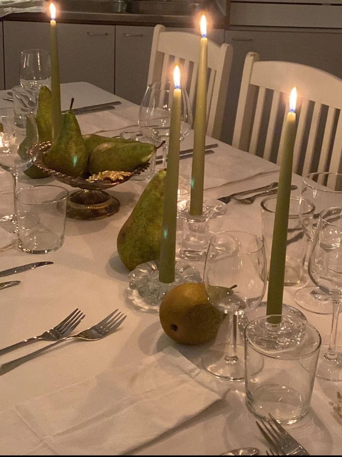 An elegant dining table set for a meal with white tablecloth, green candles, and pears as centerpiece, surrounded by glassware and silverware.