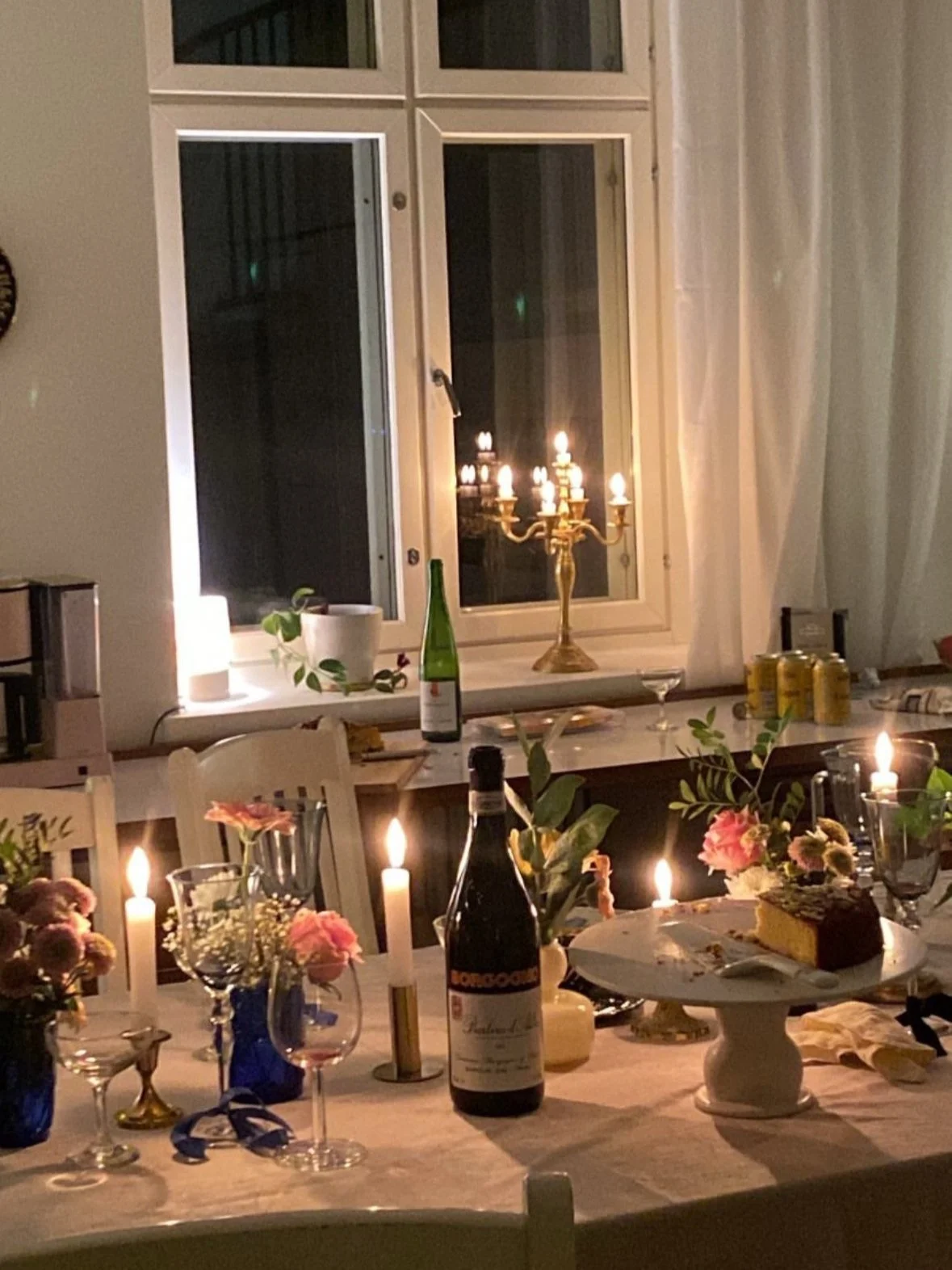Dinner table set with candles, flowers, wine, and cake in a warmly lit room with window and chandelier.