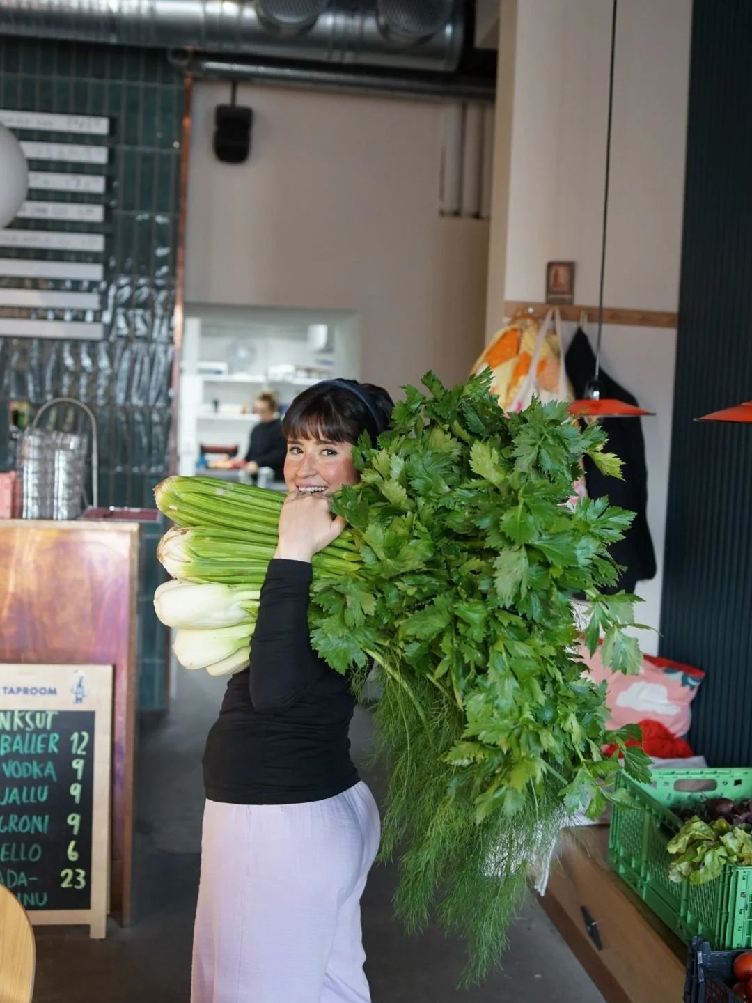 Woman smiling and holding a large bundle of fresh green vegetables, including celery and leafy greens, inside a market or grocery store.