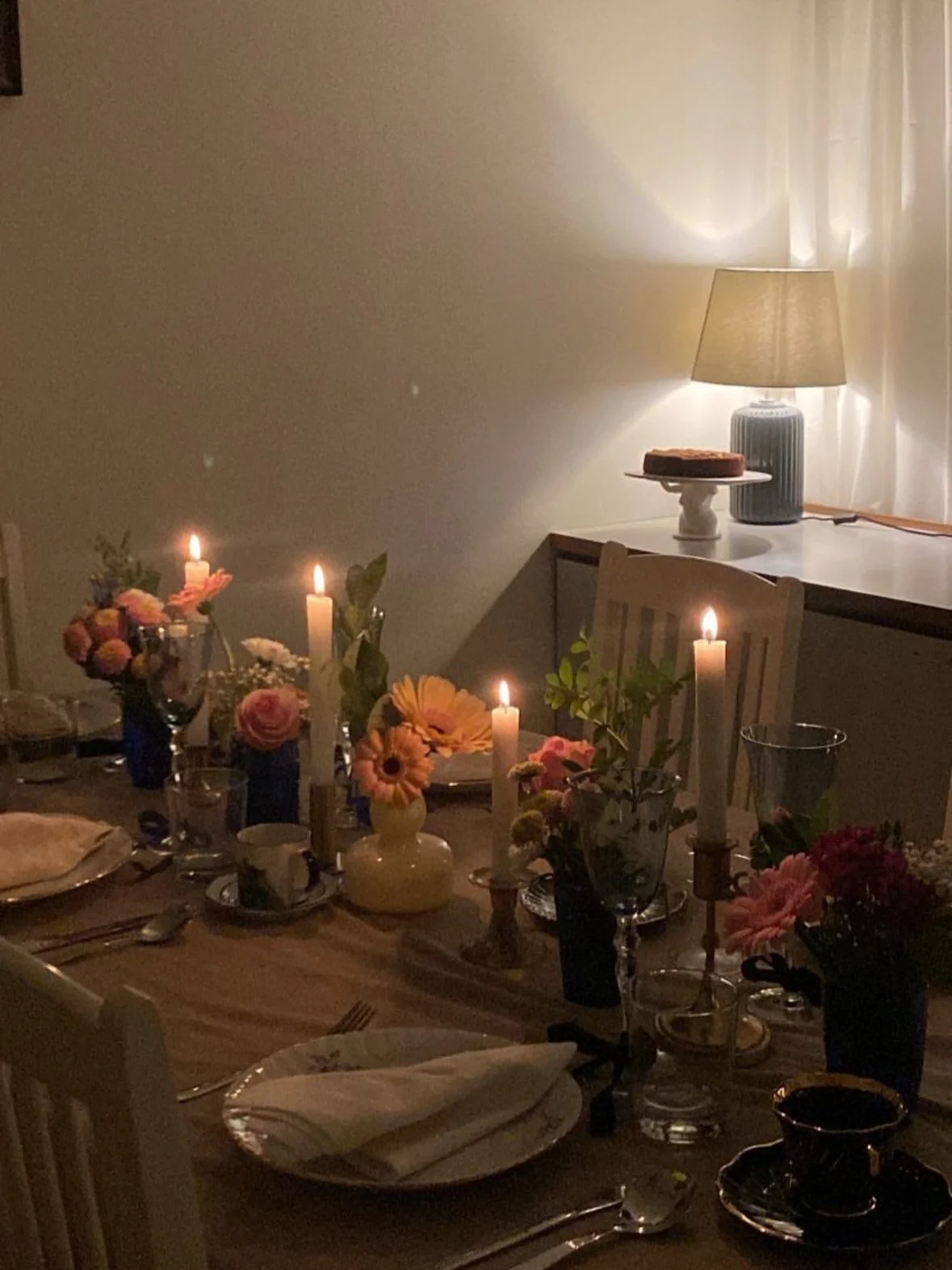 Dimly lit dining table with candles, flowers, and dishes, illuminated by a table lamp in the background.