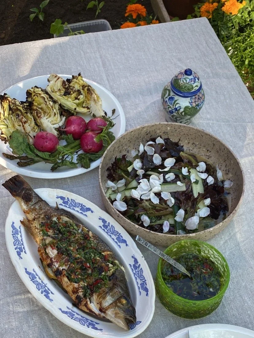 Table with grilled fish garnished with herbs and peppers, a salad with leafy greens and white edible flowers, roasted radishes, and a small green cup of dressing or sauce, set outdoors with flowers in the background.