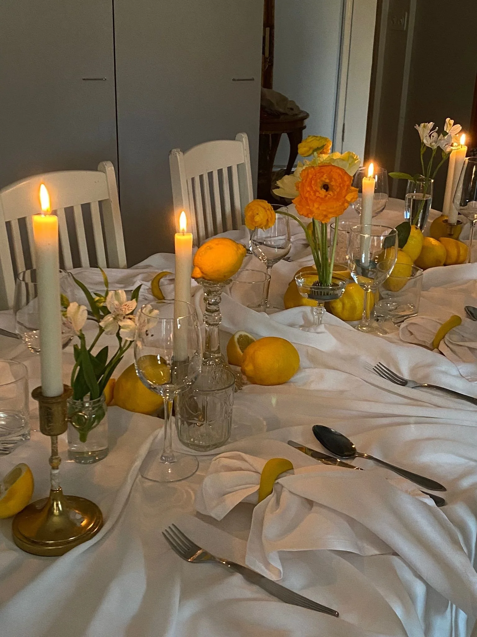 A beautifully set dining table with white tablecloth, yellow lemons, white and yellow flowers, candles, and glassware.