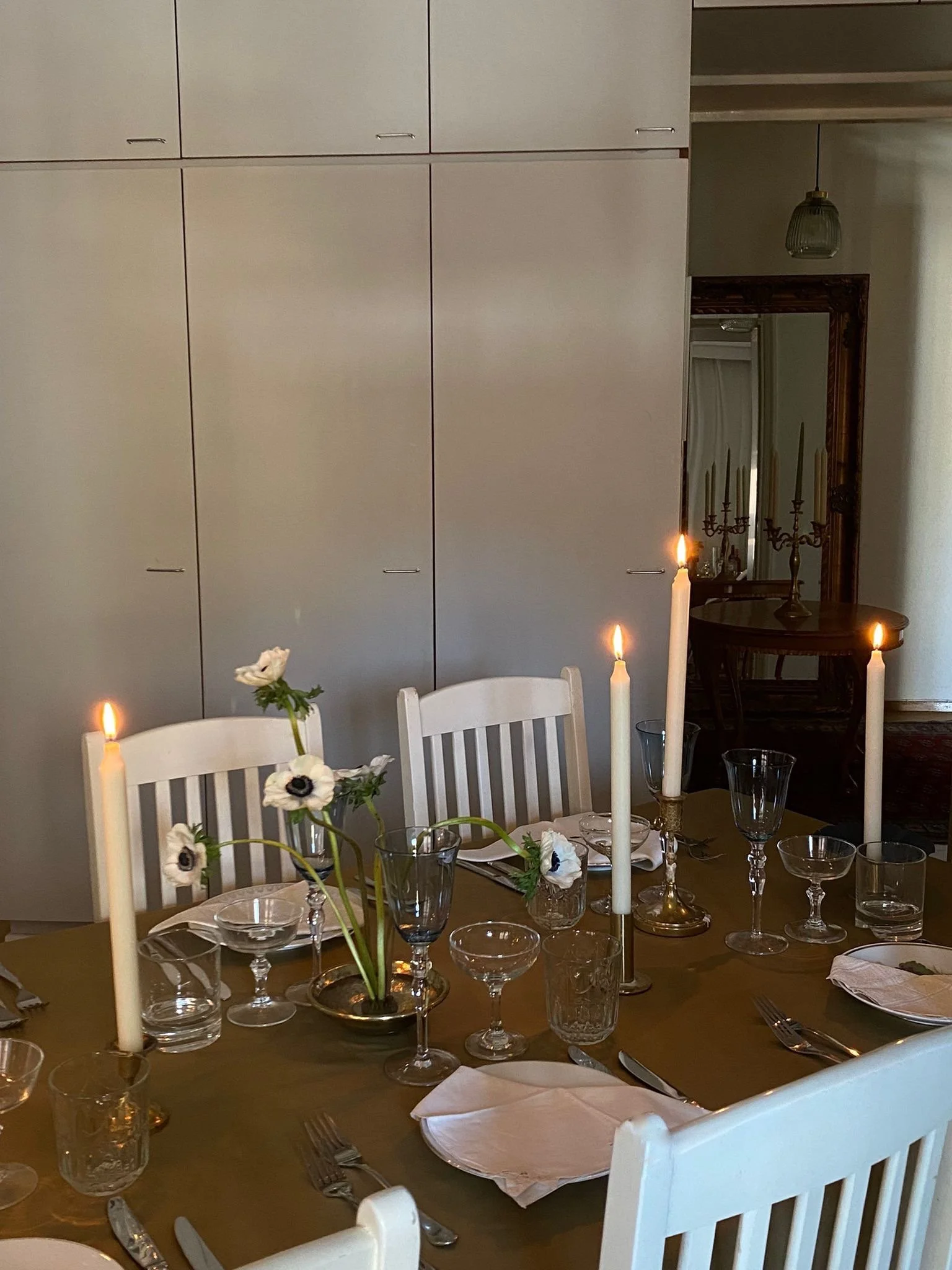 A dining table set for a meal with white chairs, candles, glassware, napkins, and a floral centerpiece with white flowers and greenery, in a room with cabinets and a mirror.
