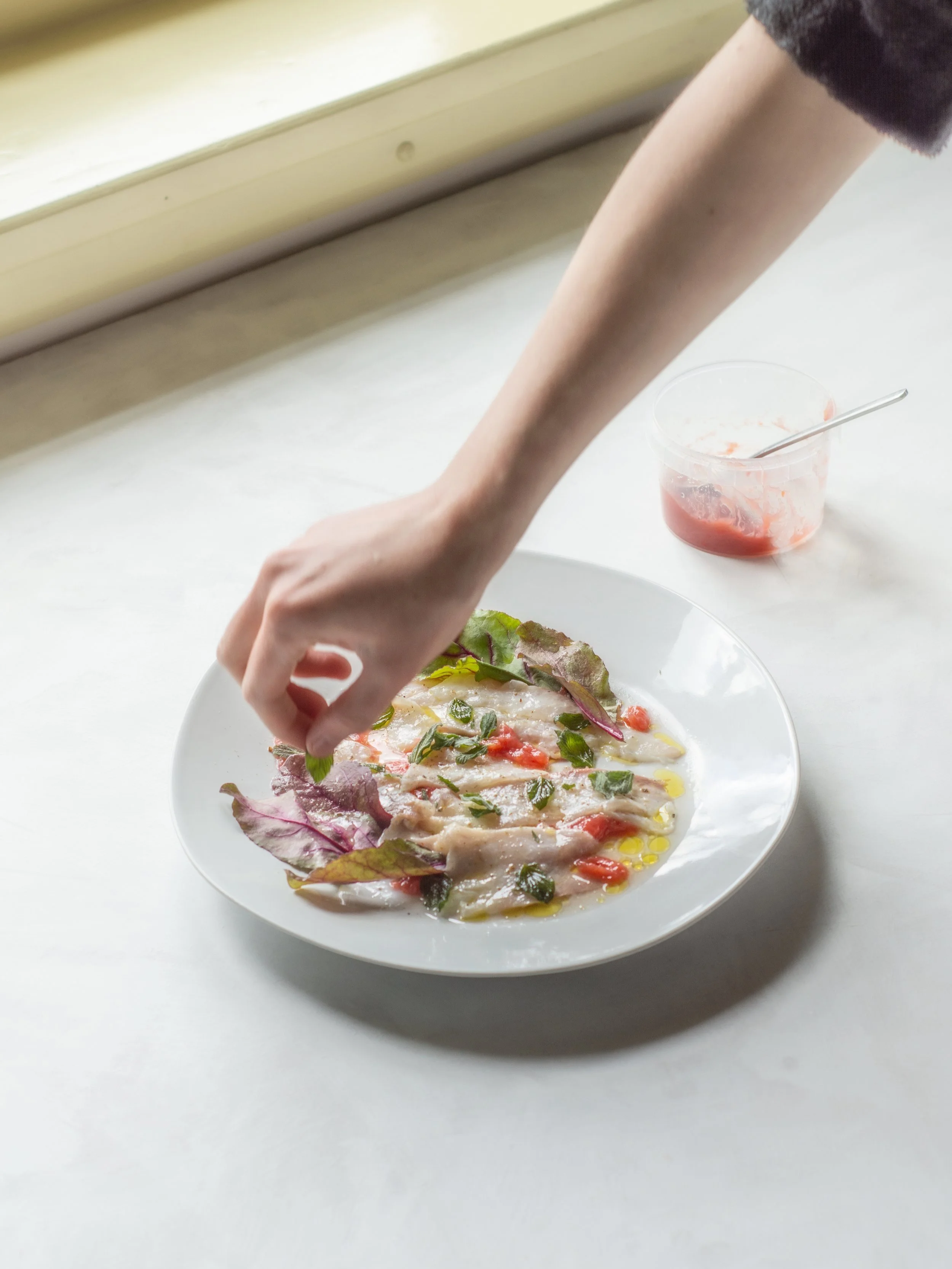 Person garnishing a plate of fish with herbs, with a small bowl of sauce nearby, on a white surface.