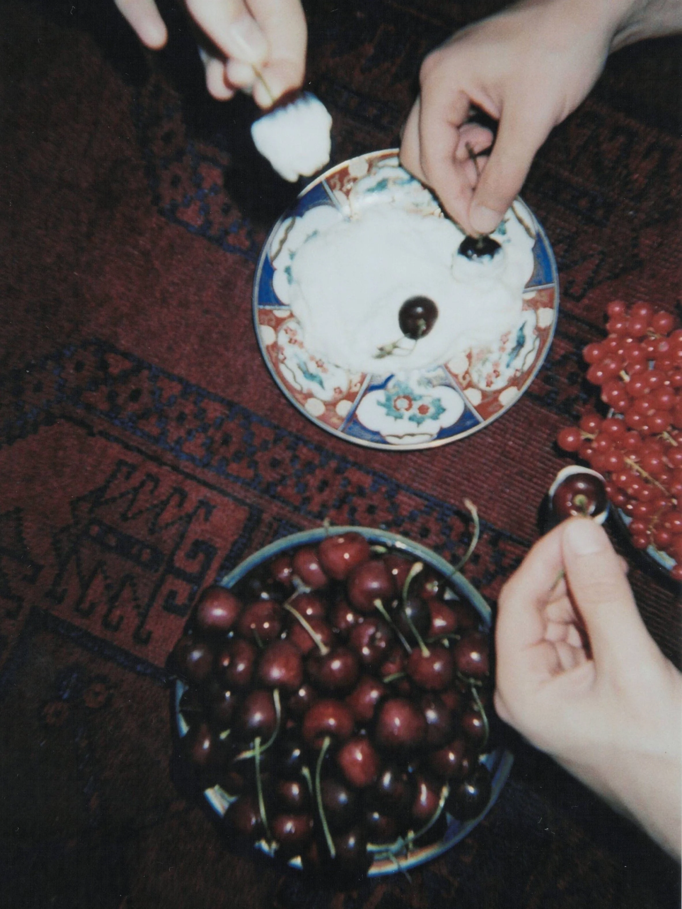 Two people are placing cherries on top of a dessert that looks like a bear's face, made with whipped cream or frosting, with cherries as eyes and nose, on a decorative plate, on a patterned rug.