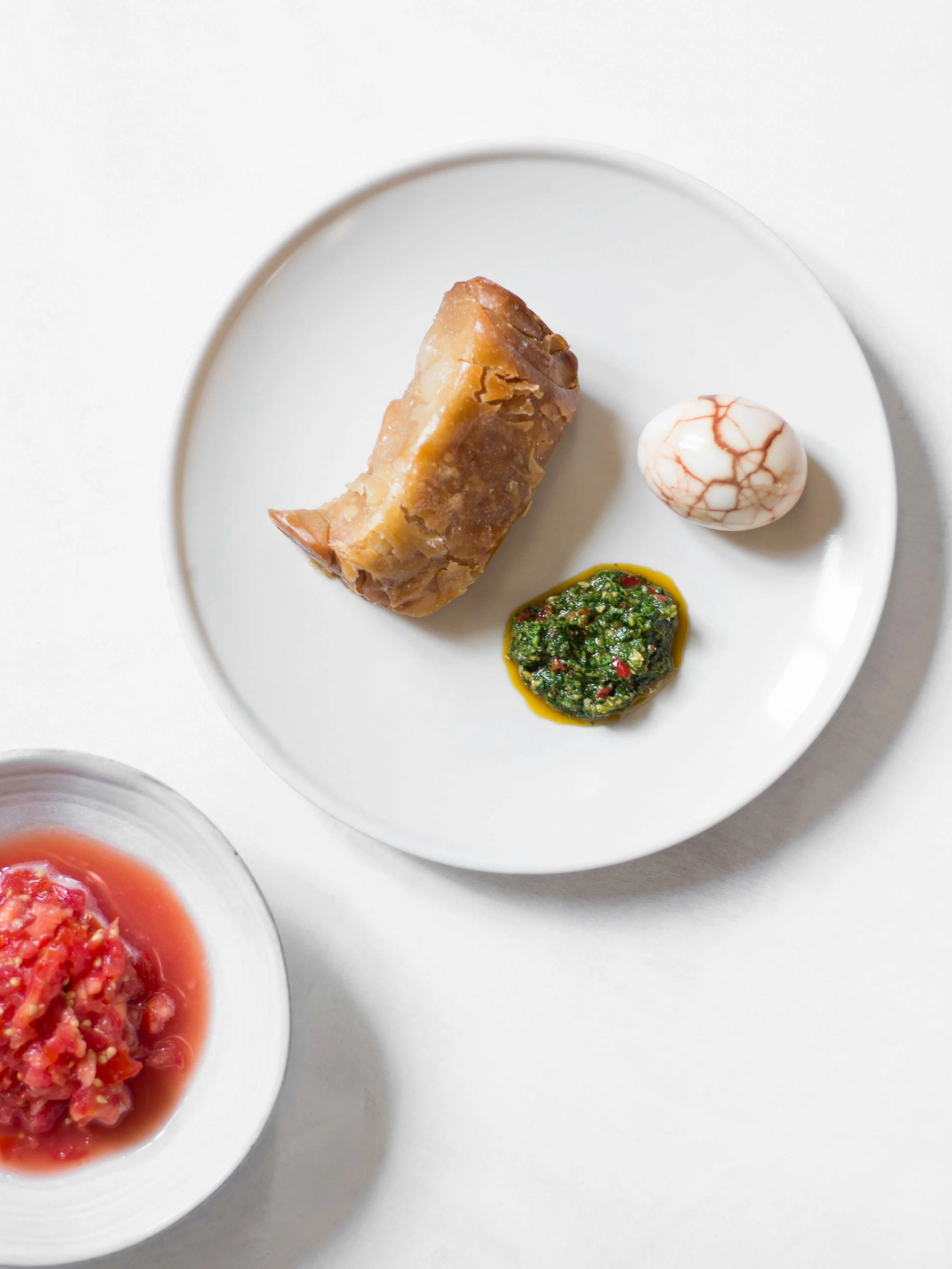 A white plate with a piece of cooked meat, a marbled boiled egg, and a dollop of green herb sauce. Part of a bowl with red tomato-based dish is visible in the bottom left corner.