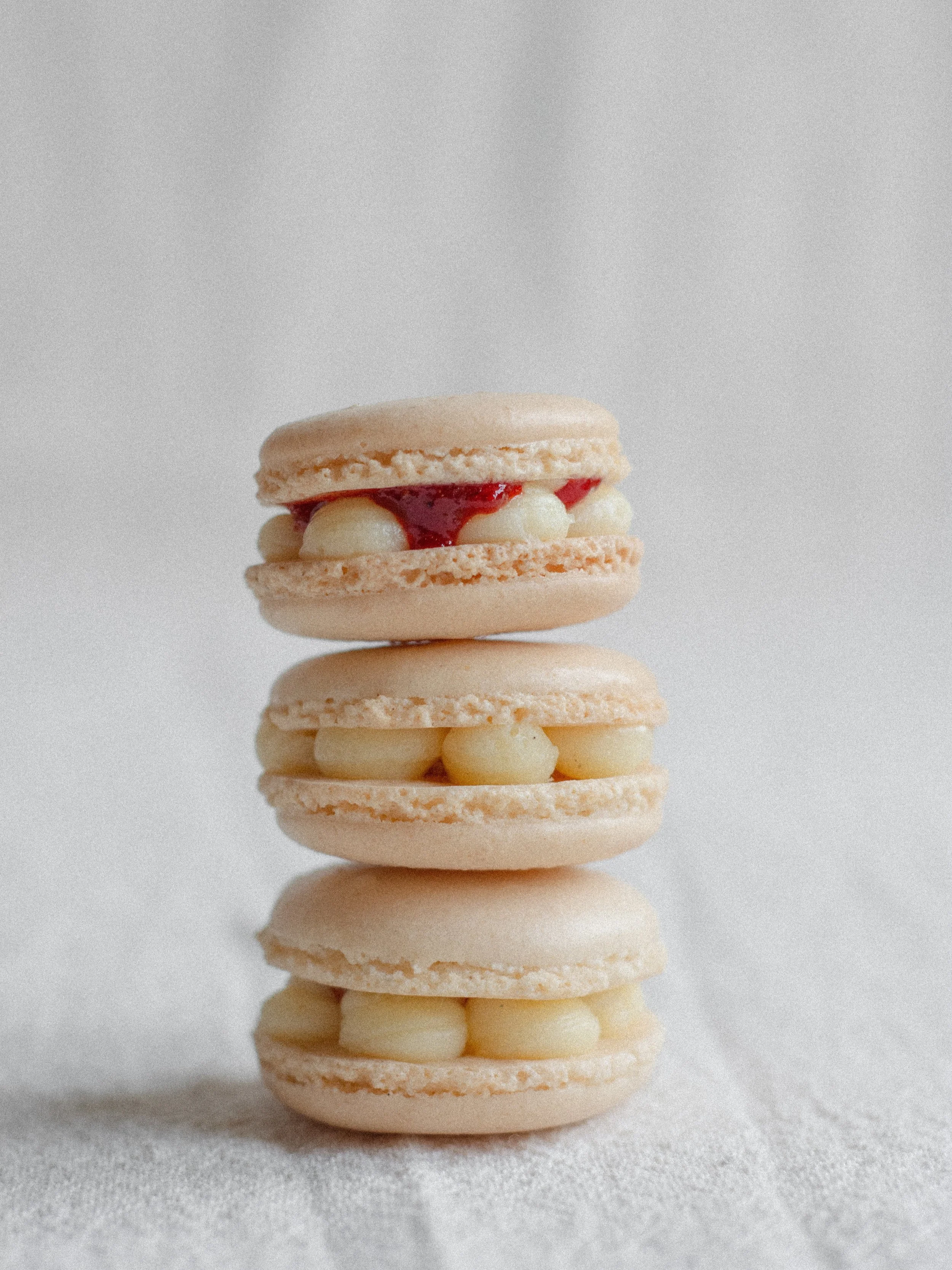 Three stacked macaron cookies with vanilla filling and some jam visible in the middle, on a light-colored surface against a plain background.