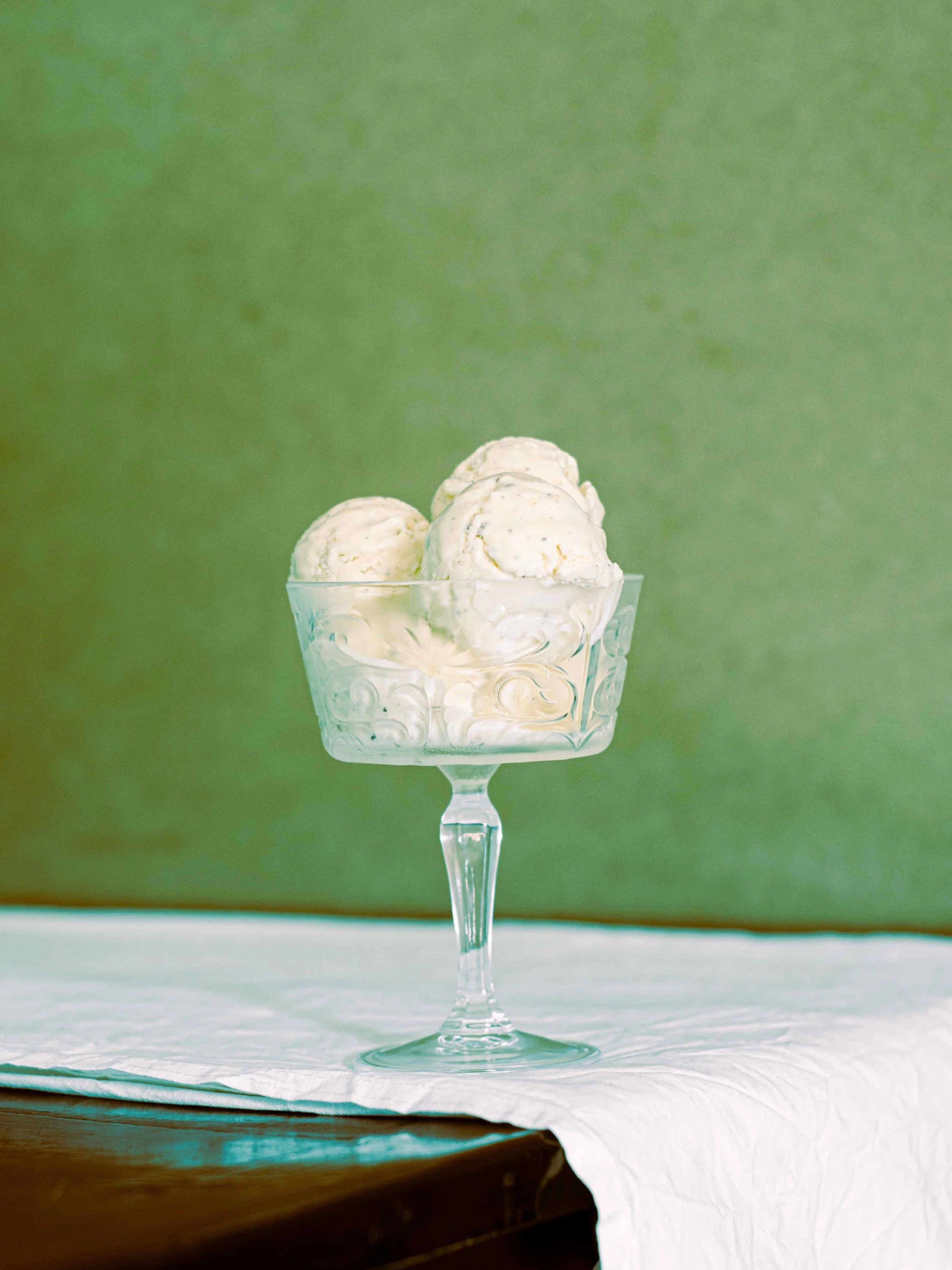Three scoops of vanilla ice cream in a decorative glass dessert cup on a white cloth-covered surface.