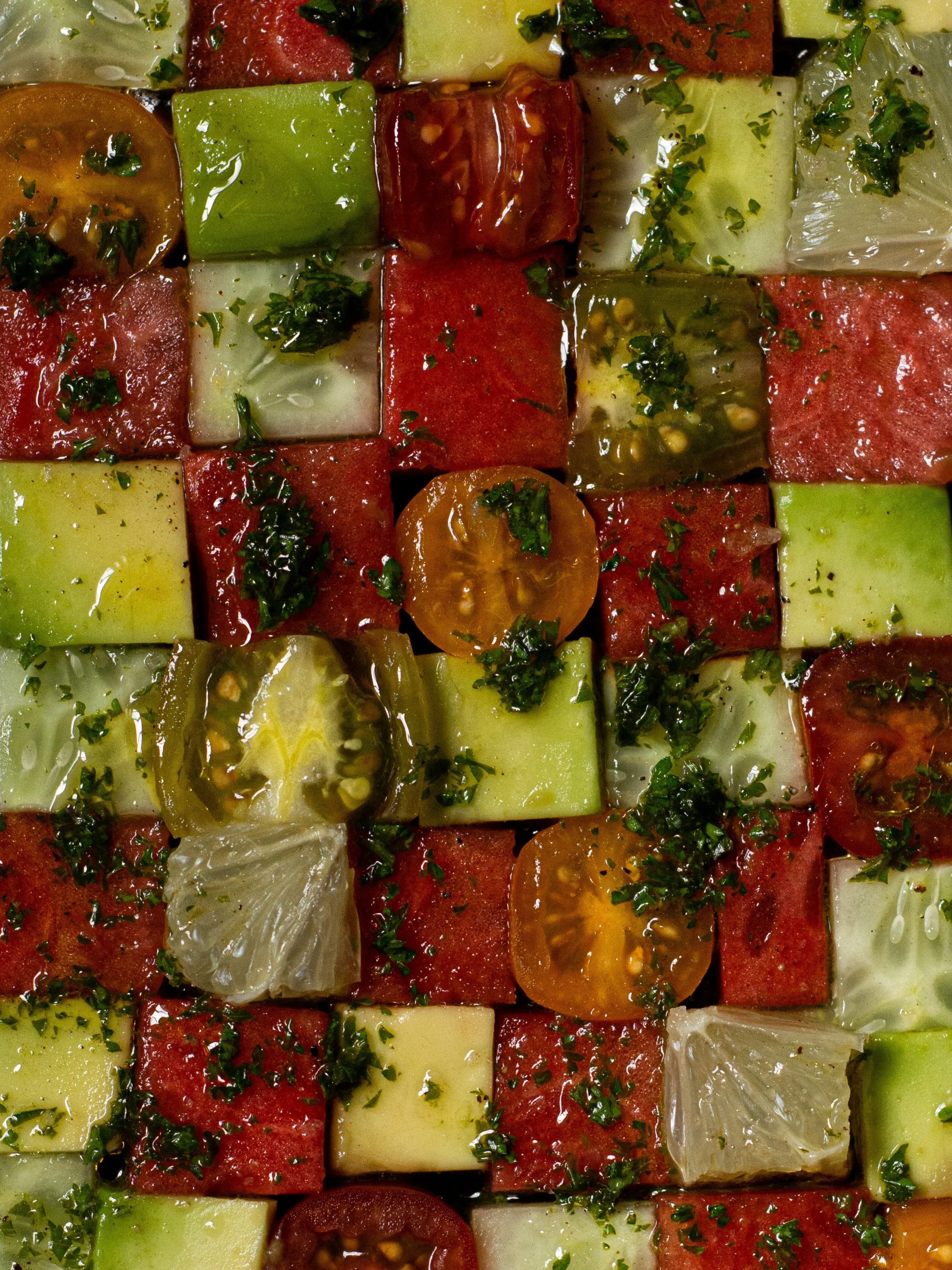 Close-up of a colorful watermelon and tomato salad with herbs, cucumbers, and lemon or lime wedges.