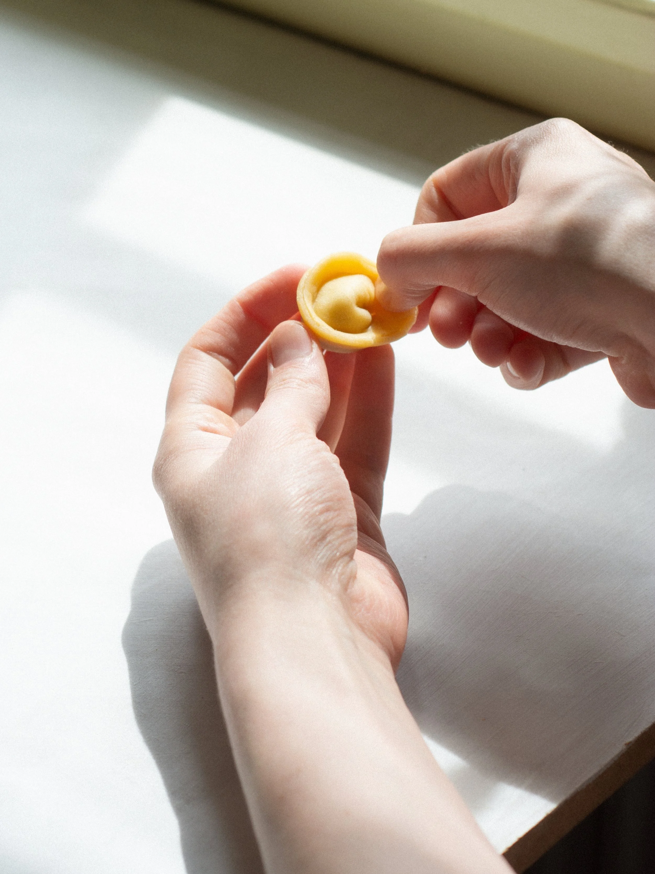 A person holding a small, round, yellow object with a heart-shaped opening, possibly a pasta or a piece of food, over a white surface by a window.