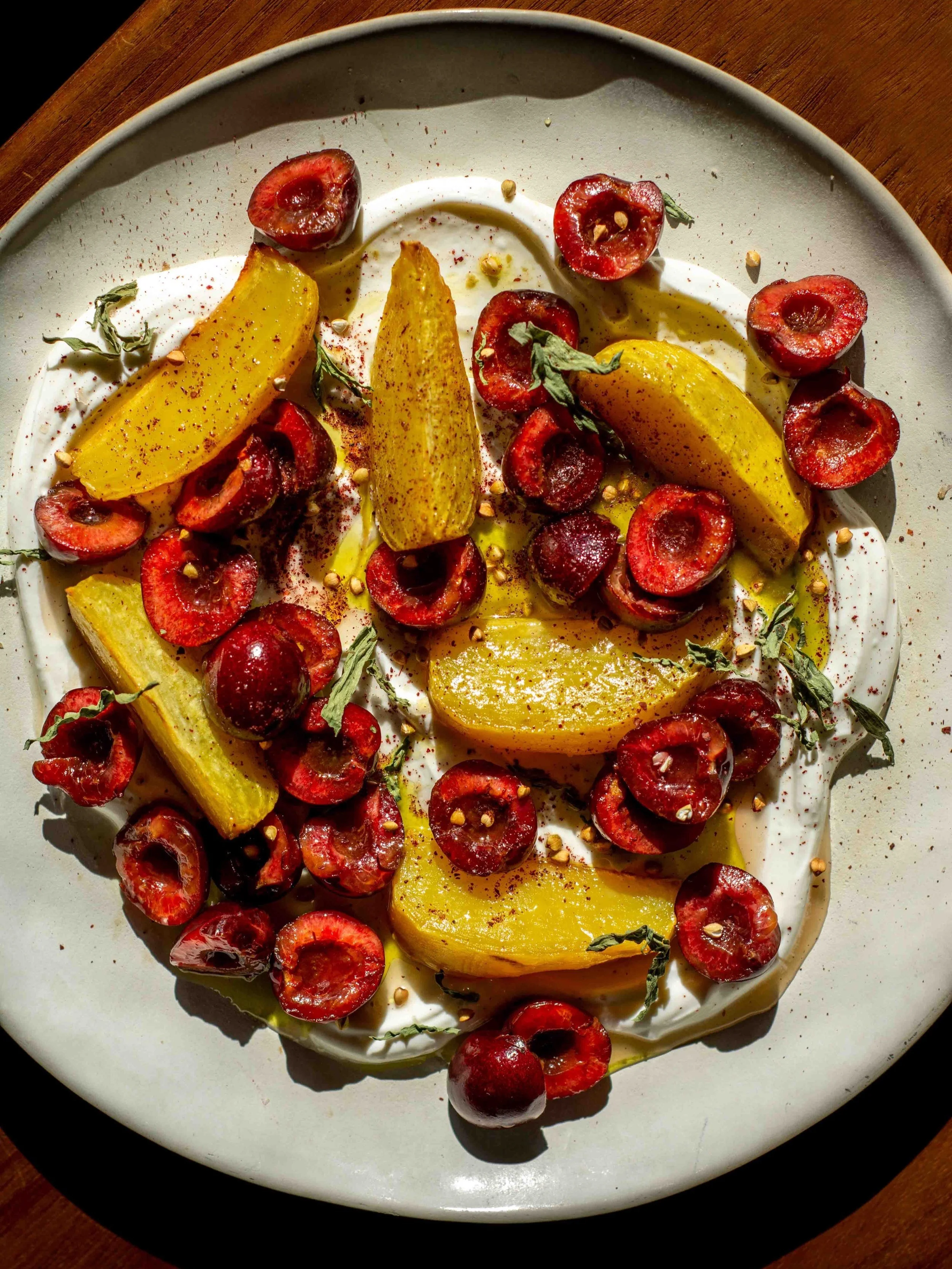 A plate of sliced yellow peaches and halved red cherries, garnished with herbs and spices, drizzled with honey