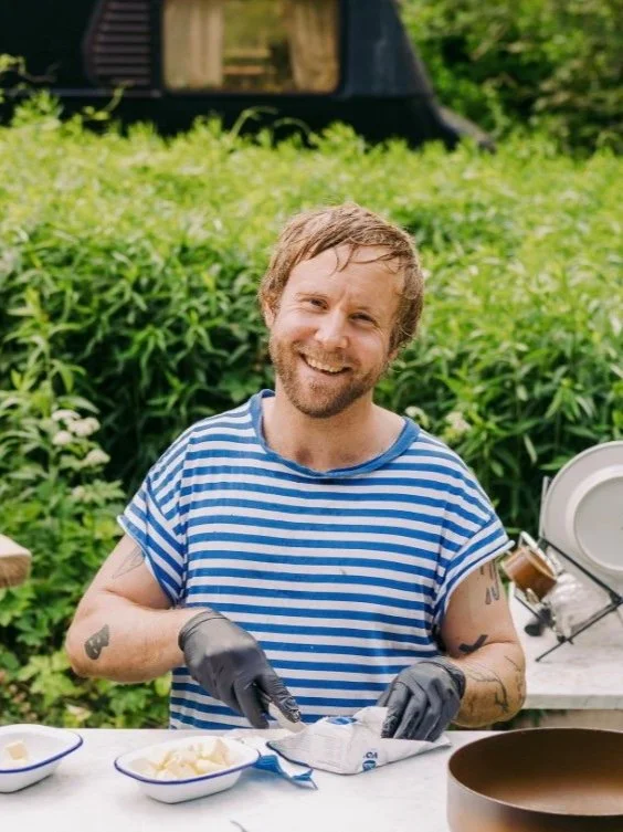 A smiling man in a blue and white striped shirt and black gloves preparing food outdoors, with greenery in the background.