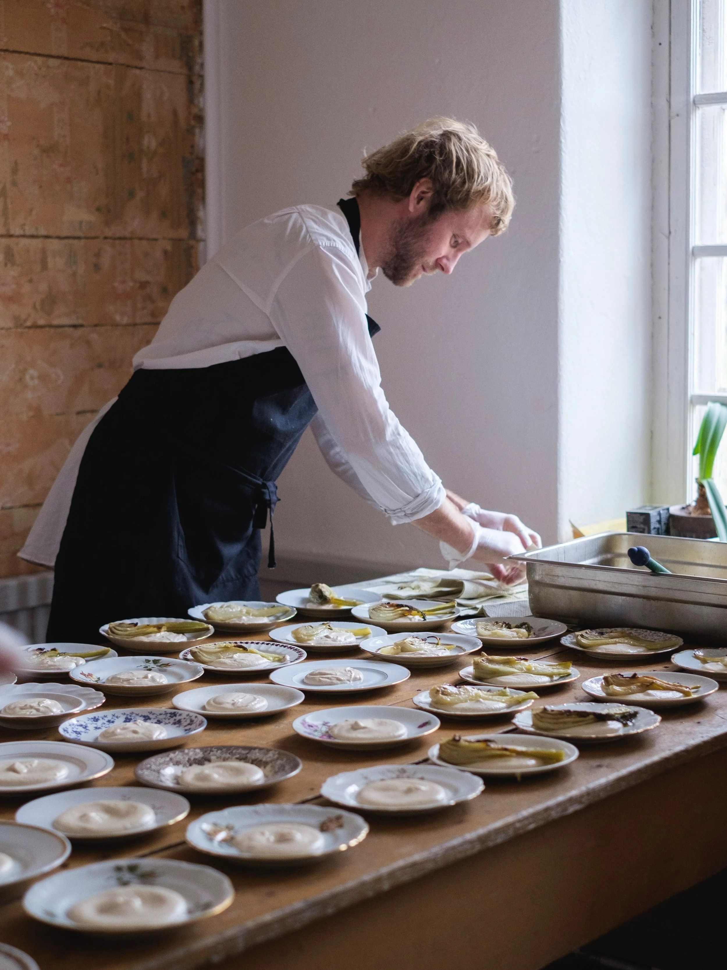 A man in a white shirt and black apron preparing plates of food on a wooden table near a window.