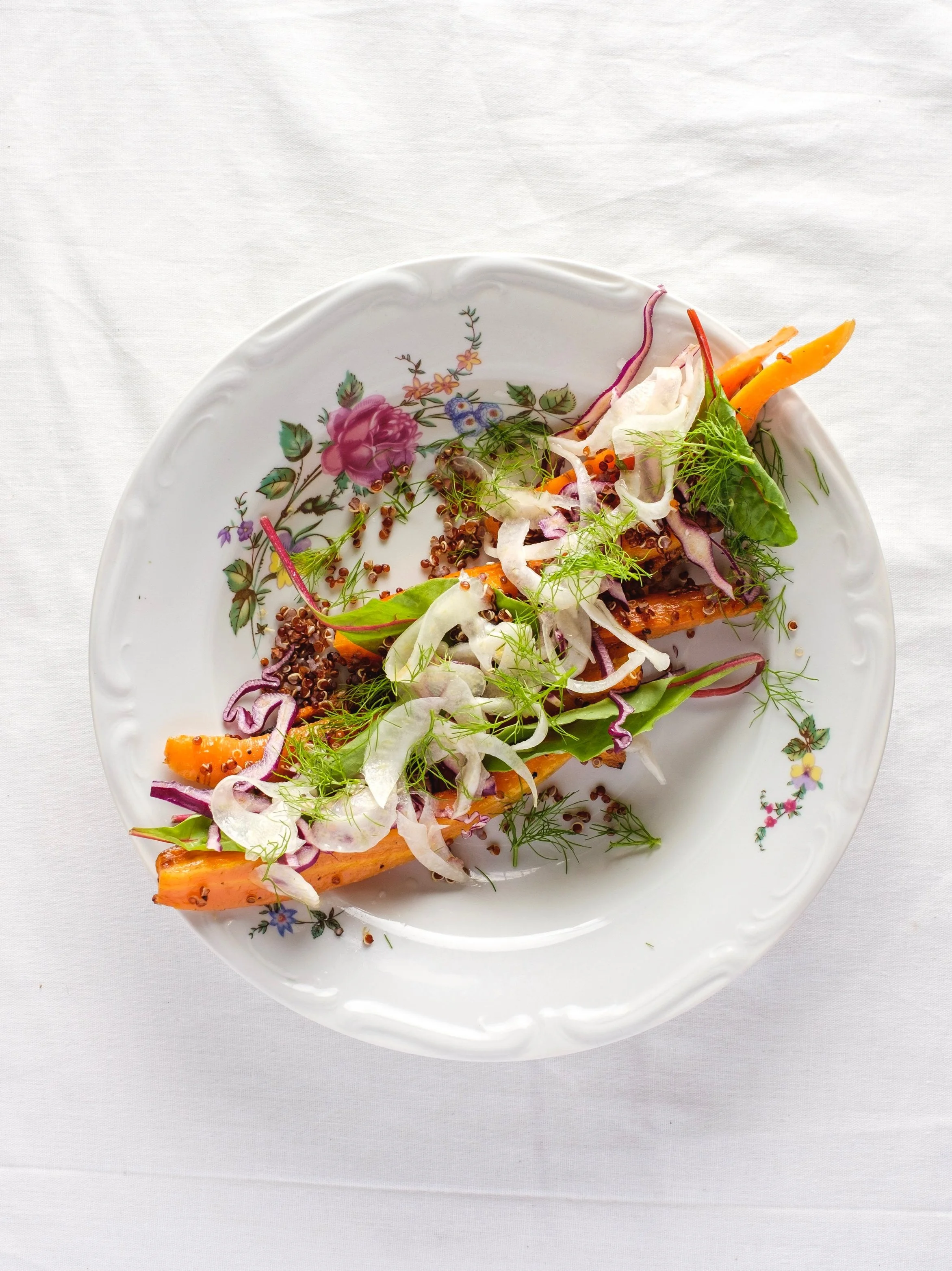A white porcelain plate with pink and green floral designs holds a fresh salad with mixed greens, shredded cabbage, carrot strips, and garnished with herbs and cracked black pepper, on a white tablecloth.