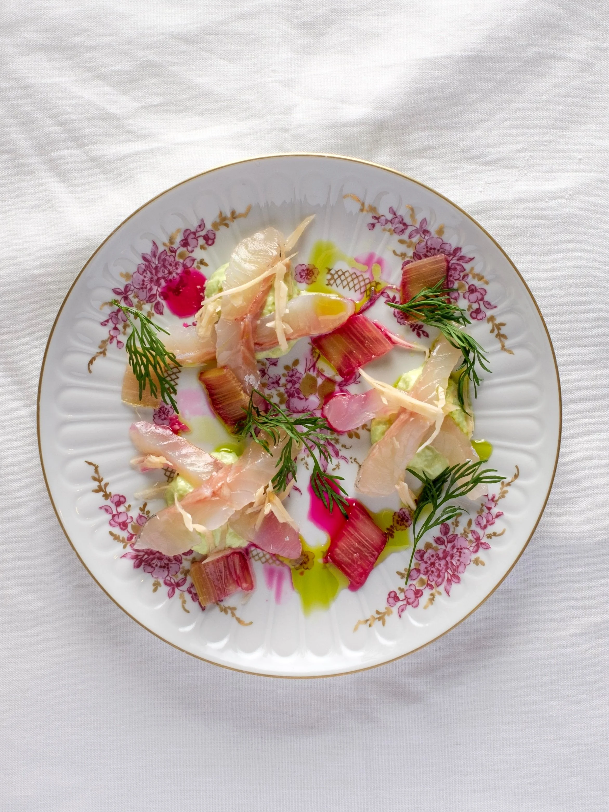 A decorative plate with finely sliced raw fish, pink and white root vegetables, fresh herbs, and drizzles of colorful oil, arranged on a white tablecloth.
