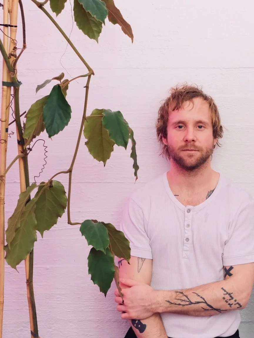A man with light brown hair and a beard, wearing a white short-sleeve Henley shirt, standing with his arms crossed, next to a leafy plant against a white wall.