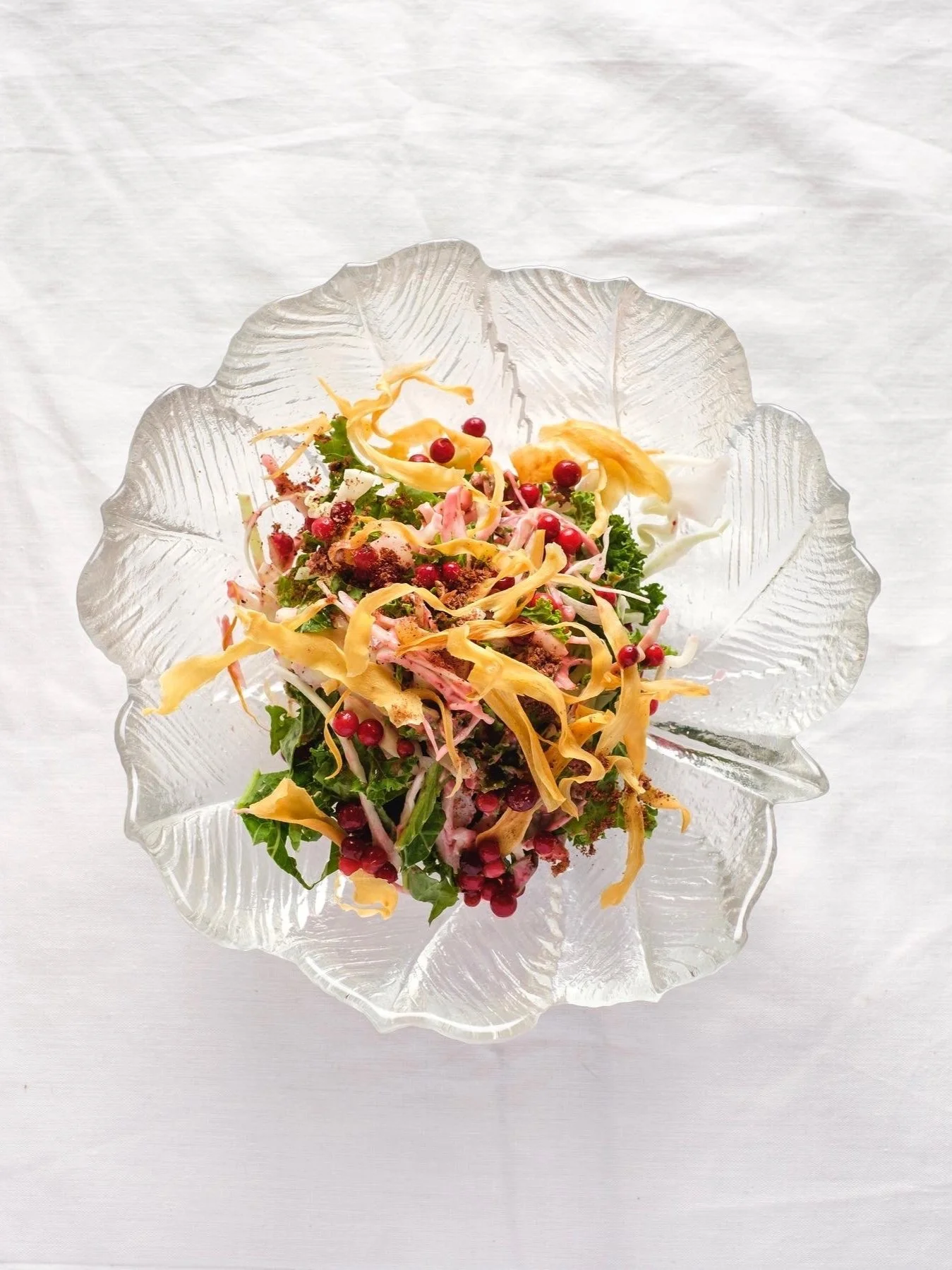 A glass bowl with a leaf-like design filled with a colorful salad containing leafy greens, shredded vegetables, red berries, and dressing on a white surface.