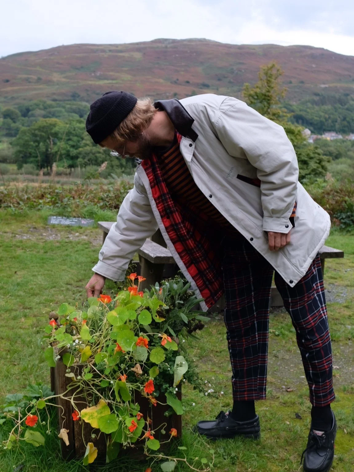 A man wearing a black beanie, glasses, a white jacket with a plaid lining, and plaid pants watering orange flowers in a garden with mountains and trees in the background.
