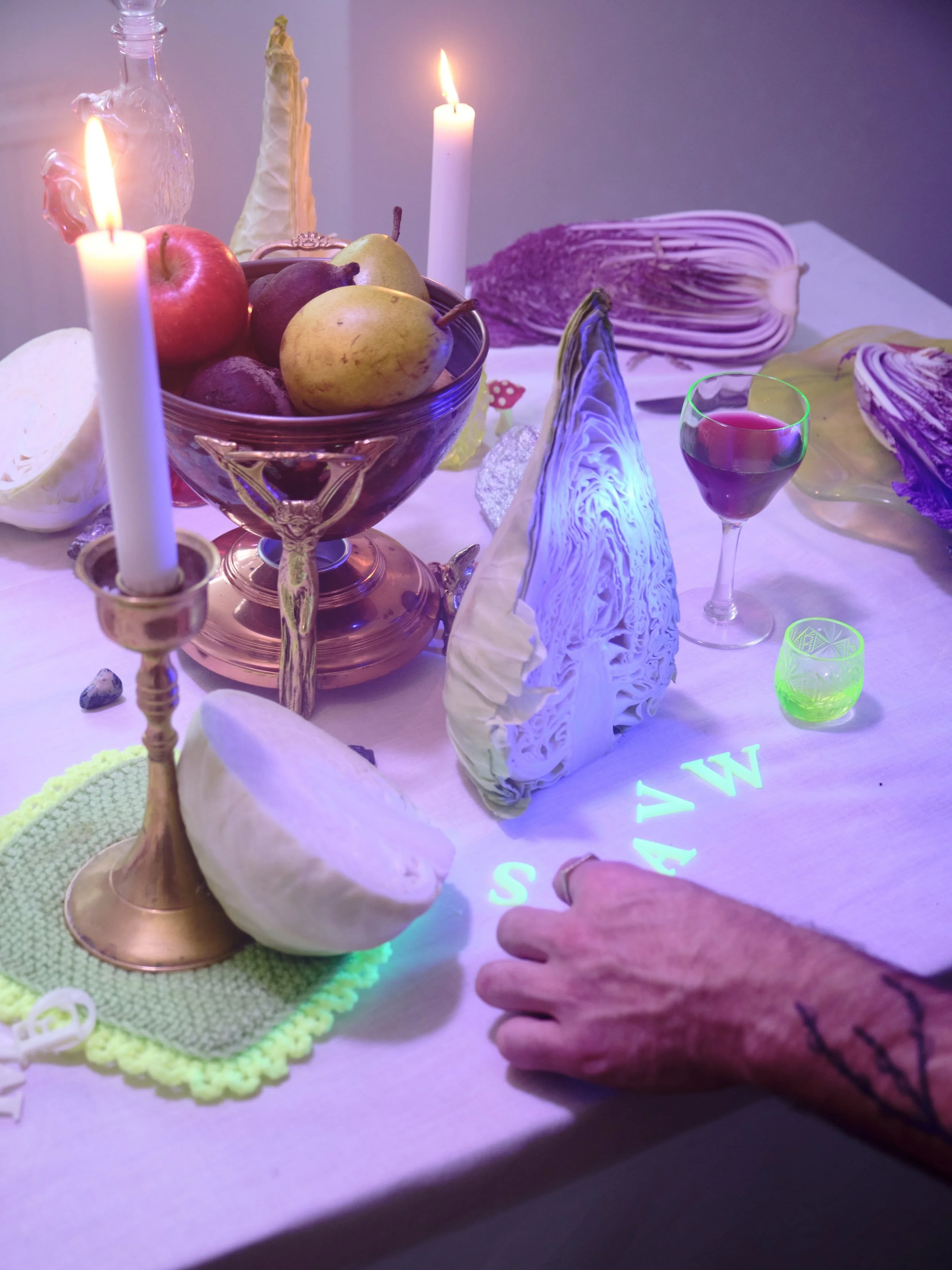A still life arrangement on a table featuring a glass bowl of assorted fruits, two lit candles, halved cabbage, colorful drinks in glasses, decorative objects, and neon lettering on the tablecloth.