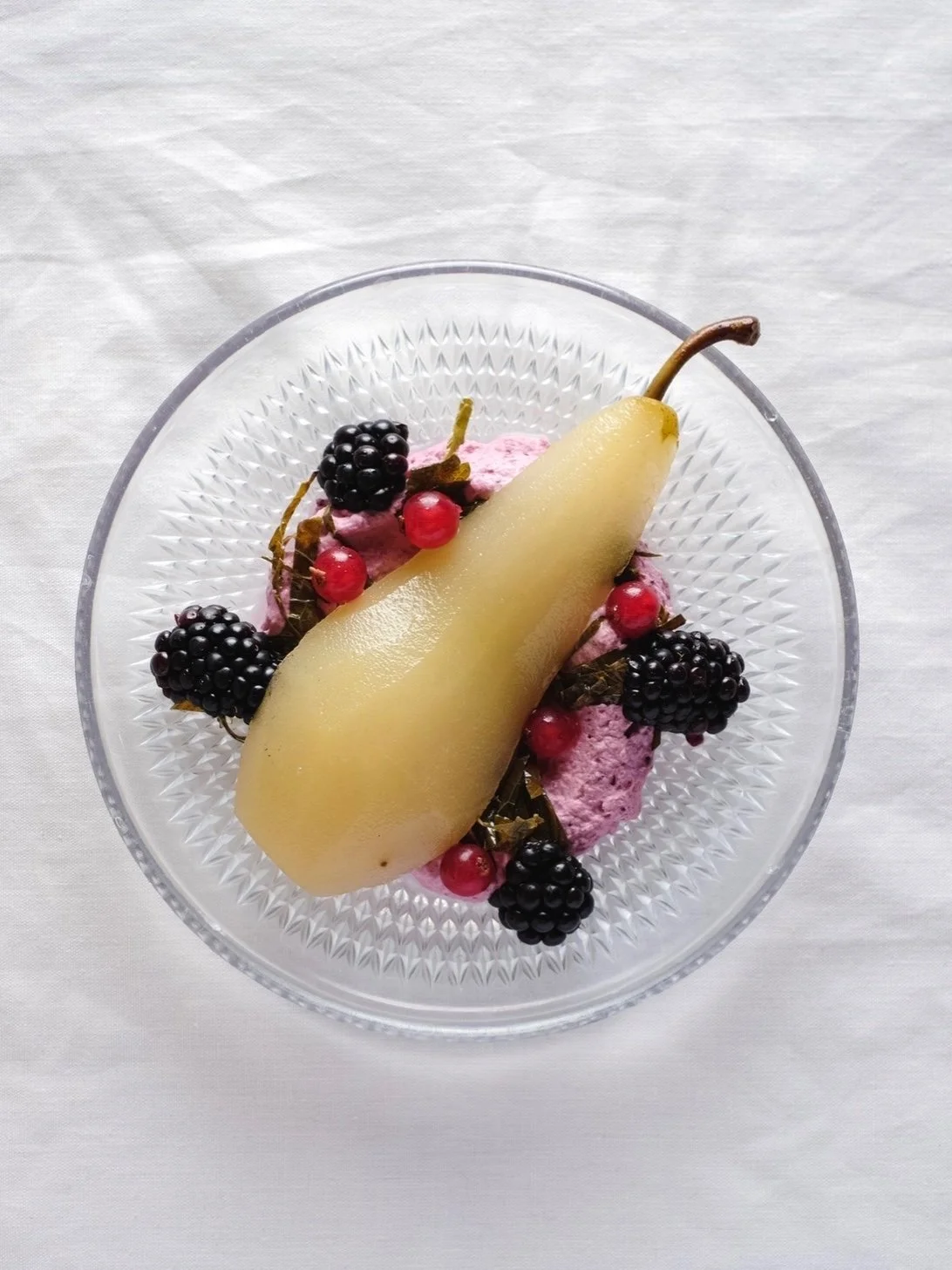 A glass bowl containing a poached pear, blackberries, red currants, and pink ice cream at the center, on a white tablecloth.