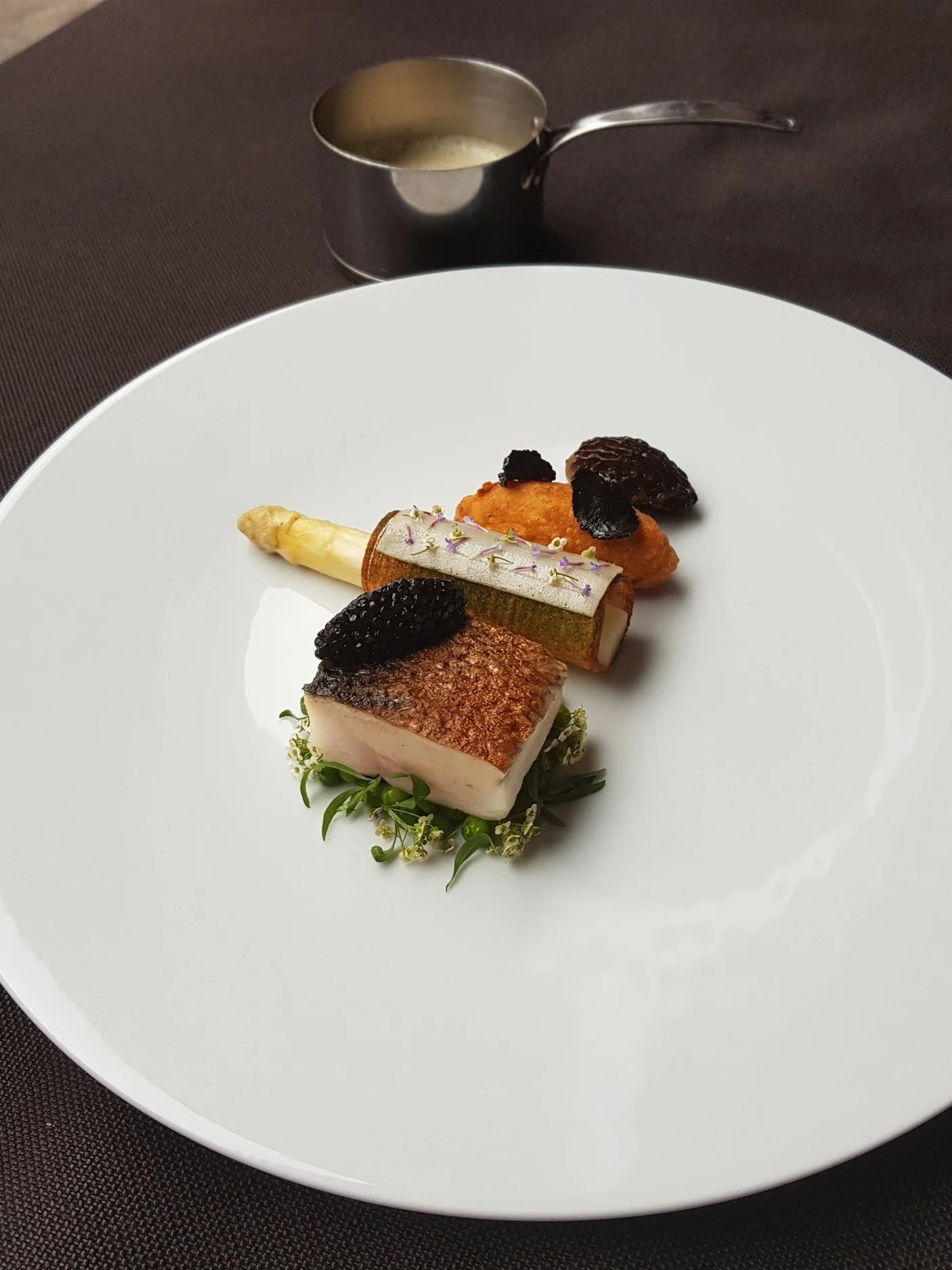 A fine dining plate with a piece of seared fish, a charred vegetable, and topped with black caviar, garnished with herbs and edible flowers. In the background, a small metal pitcher with sauce, on a dark tablecloth.