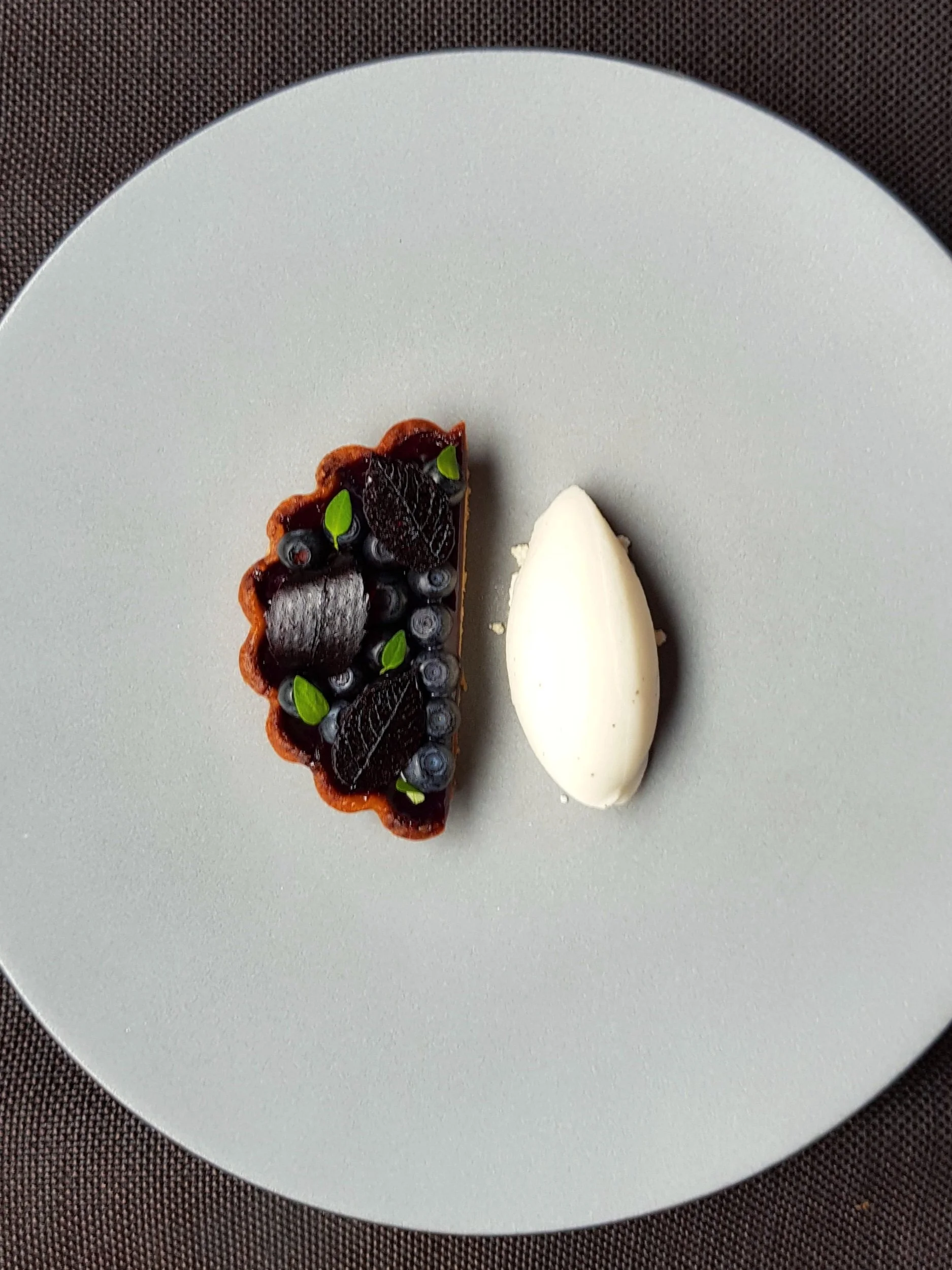 A dessert plate with a small tart topped with blueberries, blackberries, and green leaves, and a dollop of white cream or yogurt on the side.